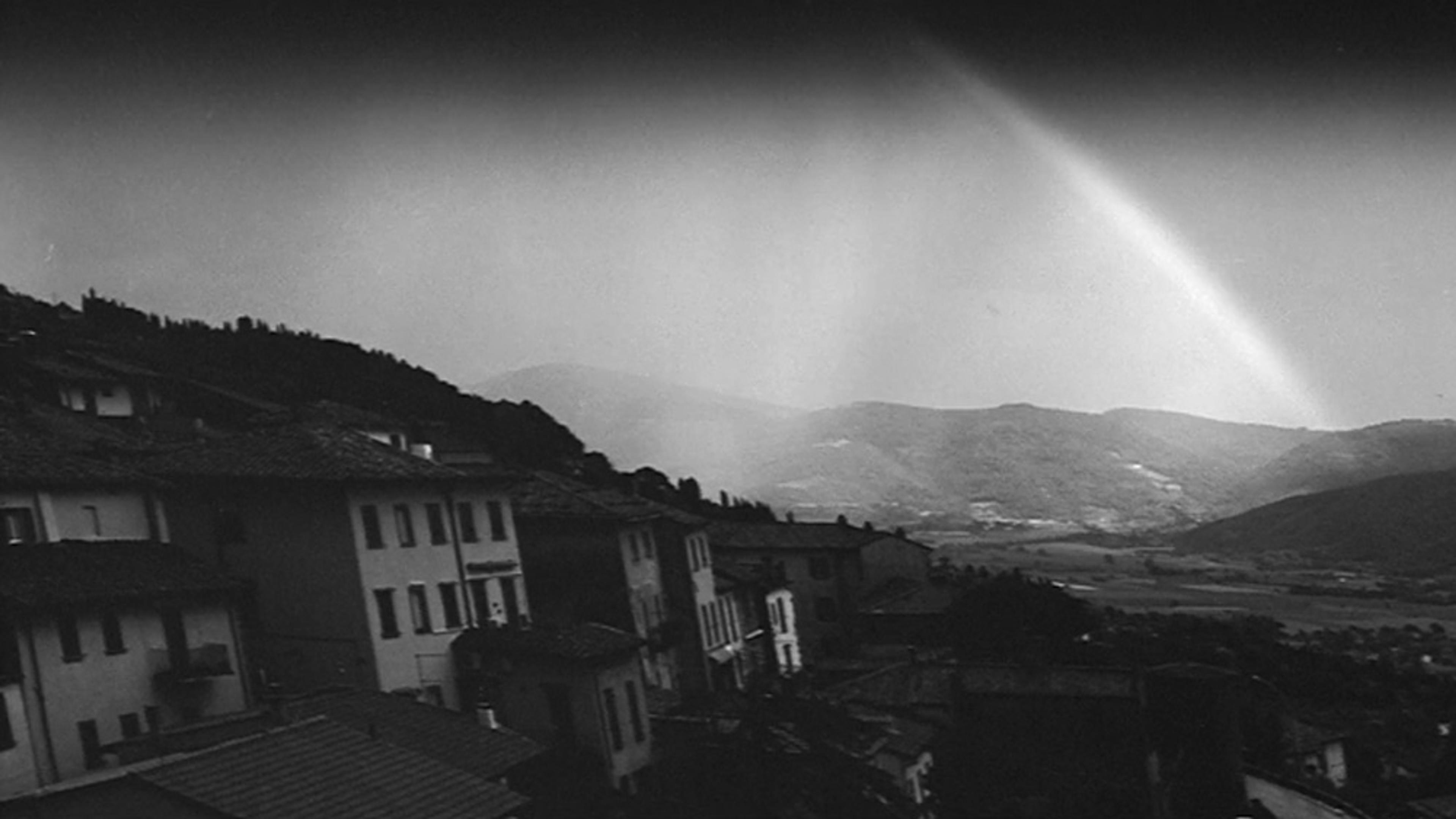 Black and white photo of hillside houses with a rainbow arcing over distant mountains and a valley.