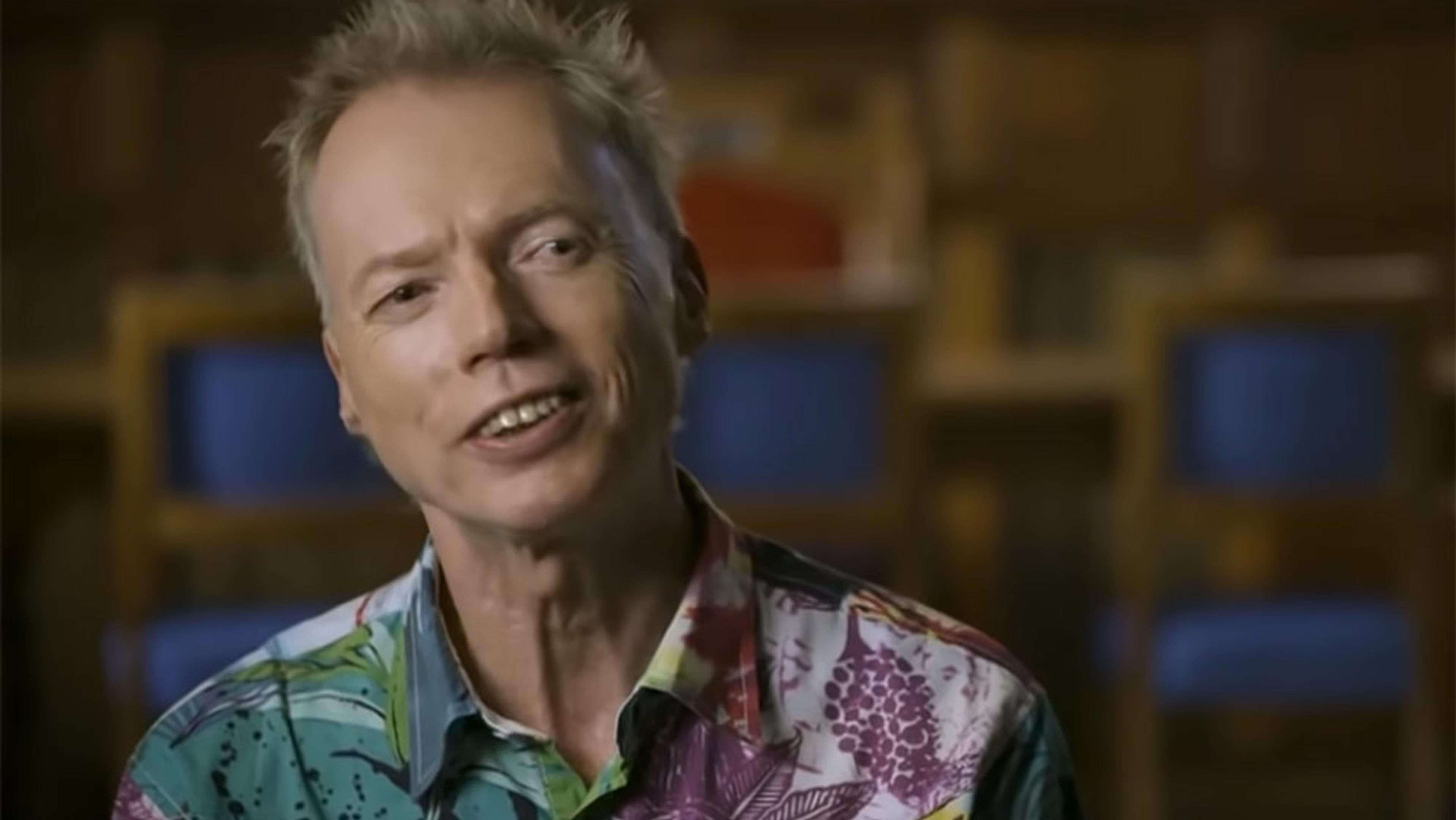 A man in a colourful shirt smiles in a photo. The background is blurred, showing blue chairs and wooden shelves, suggesting a library.