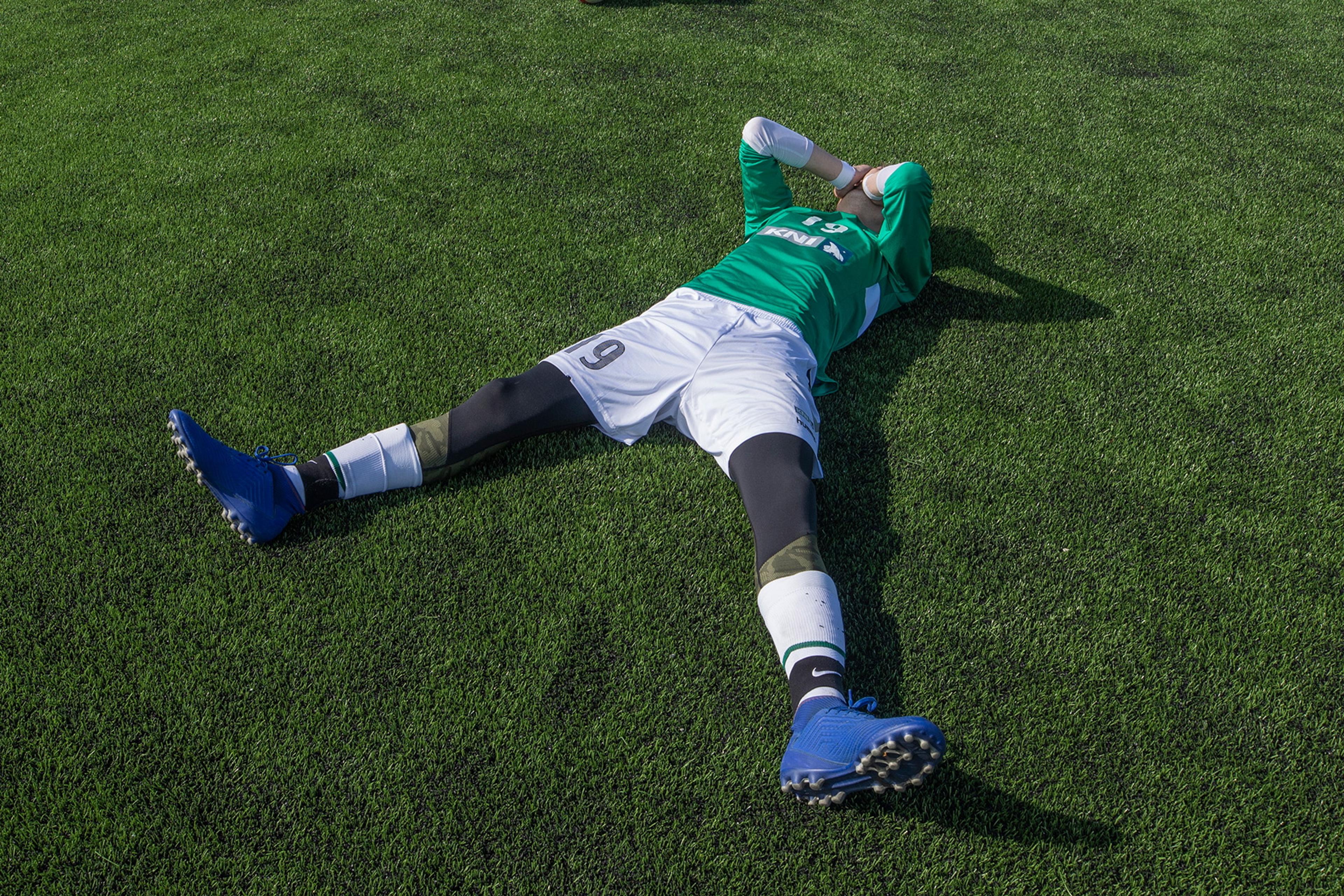 Photo of a footballer lying on a pitch looking distressed with a child nearby holding a ball.