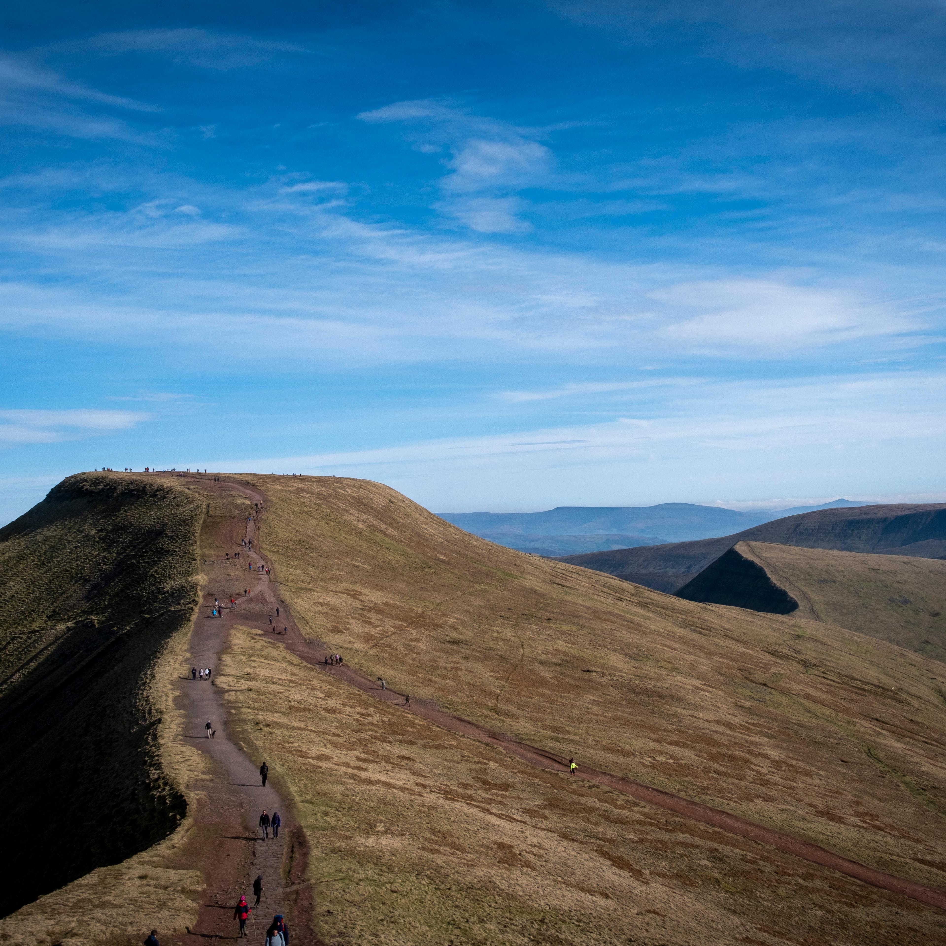 A mountain peak with a walking path and hikers, under a clear blue sky with scattered clouds.