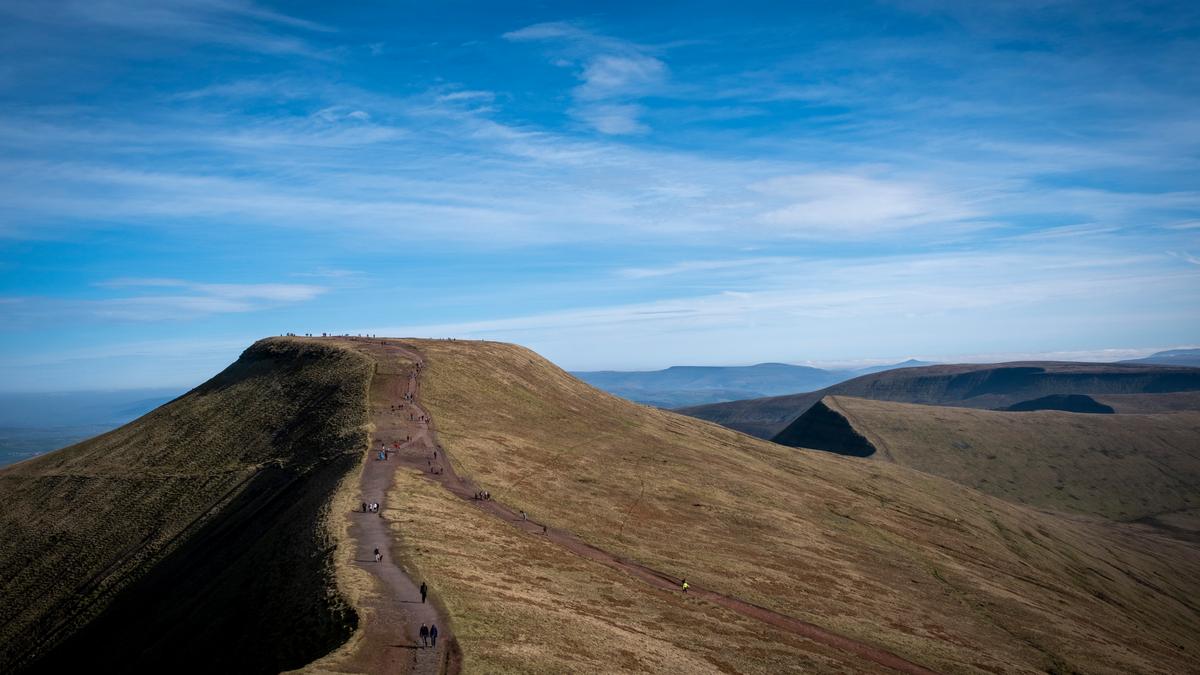 A mountain peak with a walking path and hikers, under a clear blue sky with scattered clouds.