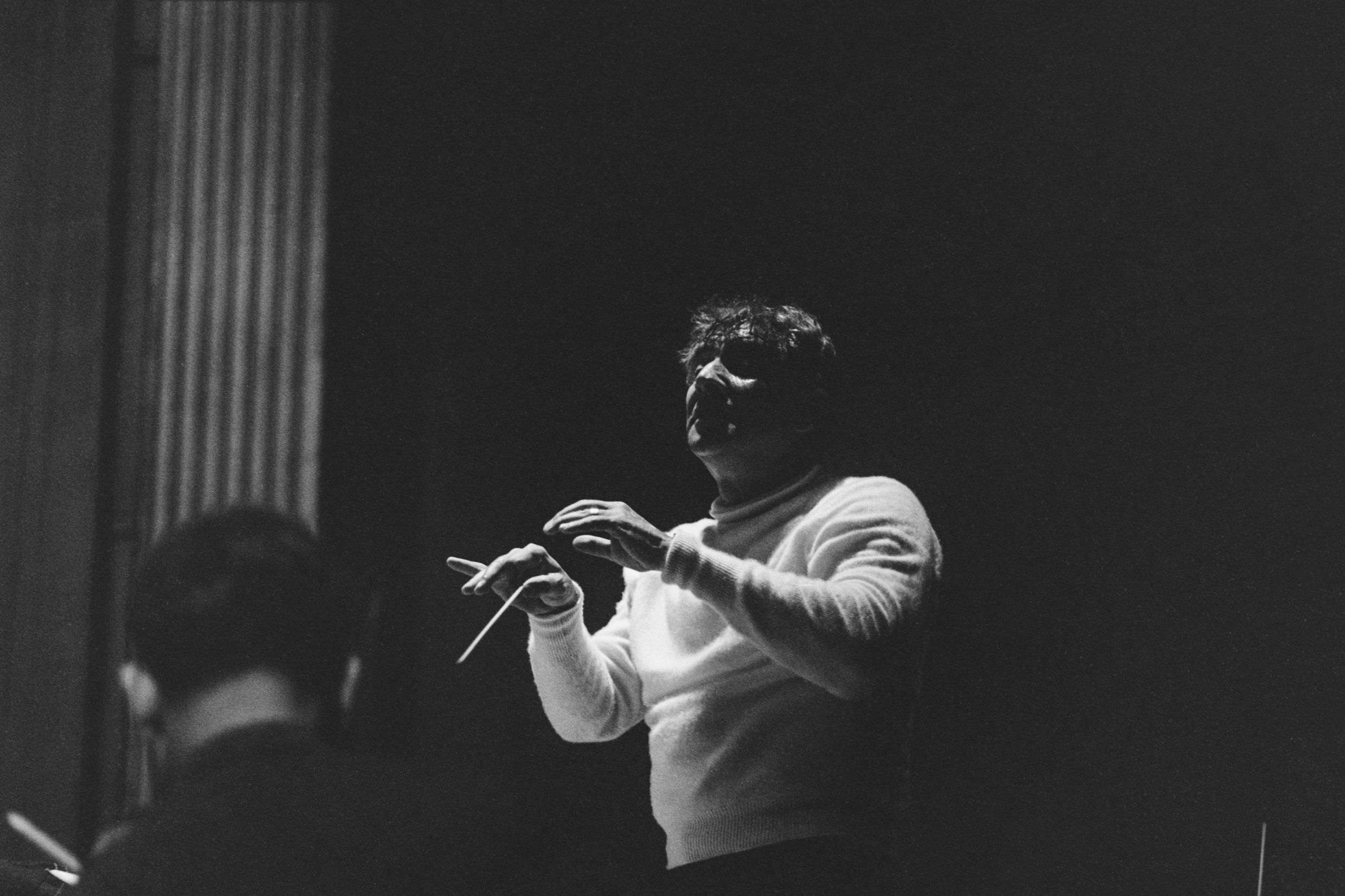 Black and white photo of a conductor in a white jumper gesturing with a baton, partially visible orchestra member in foreground.