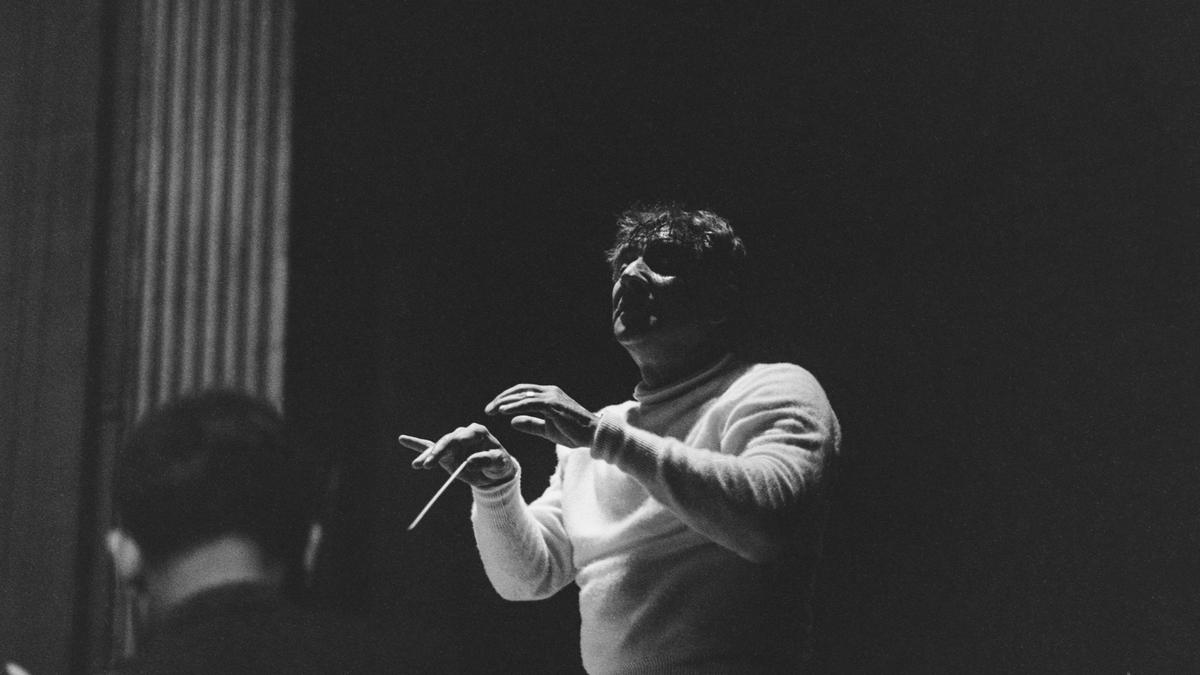 Black and white photo of a conductor in a white jumper gesturing with a baton, partially visible orchestra member in foreground.