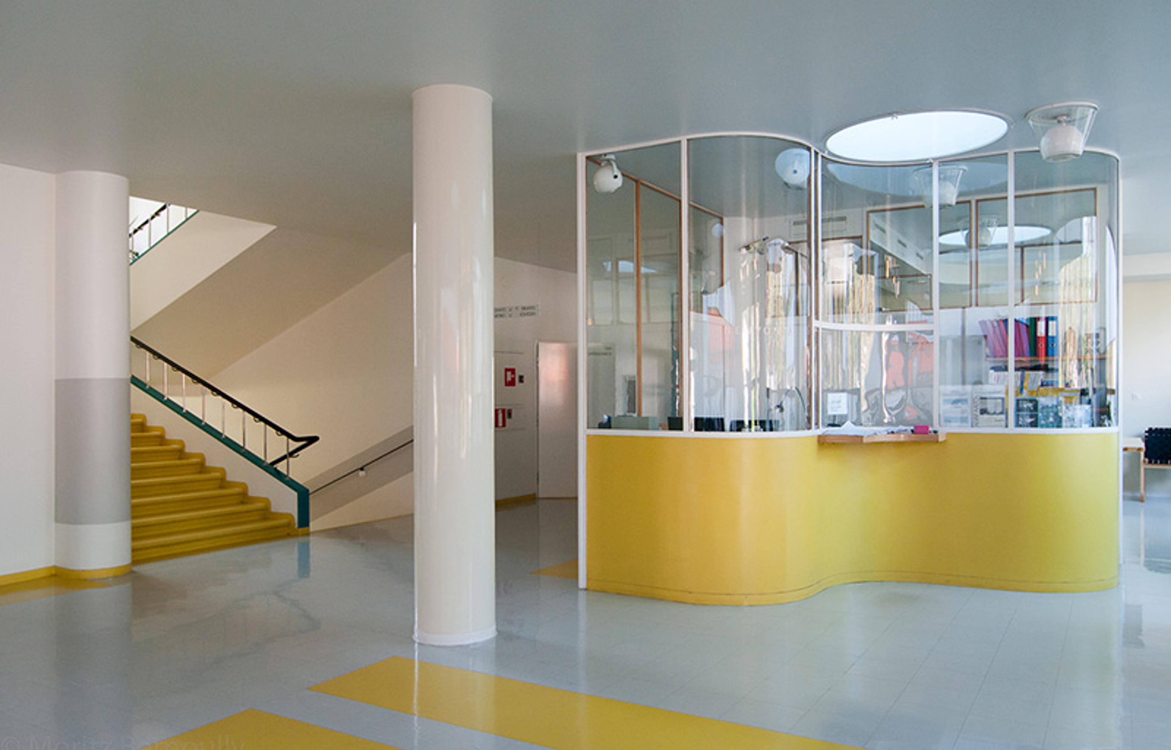A modern interior with yellow accents, a staircase on the left, and a round reception desk enclosed in glass on the right.