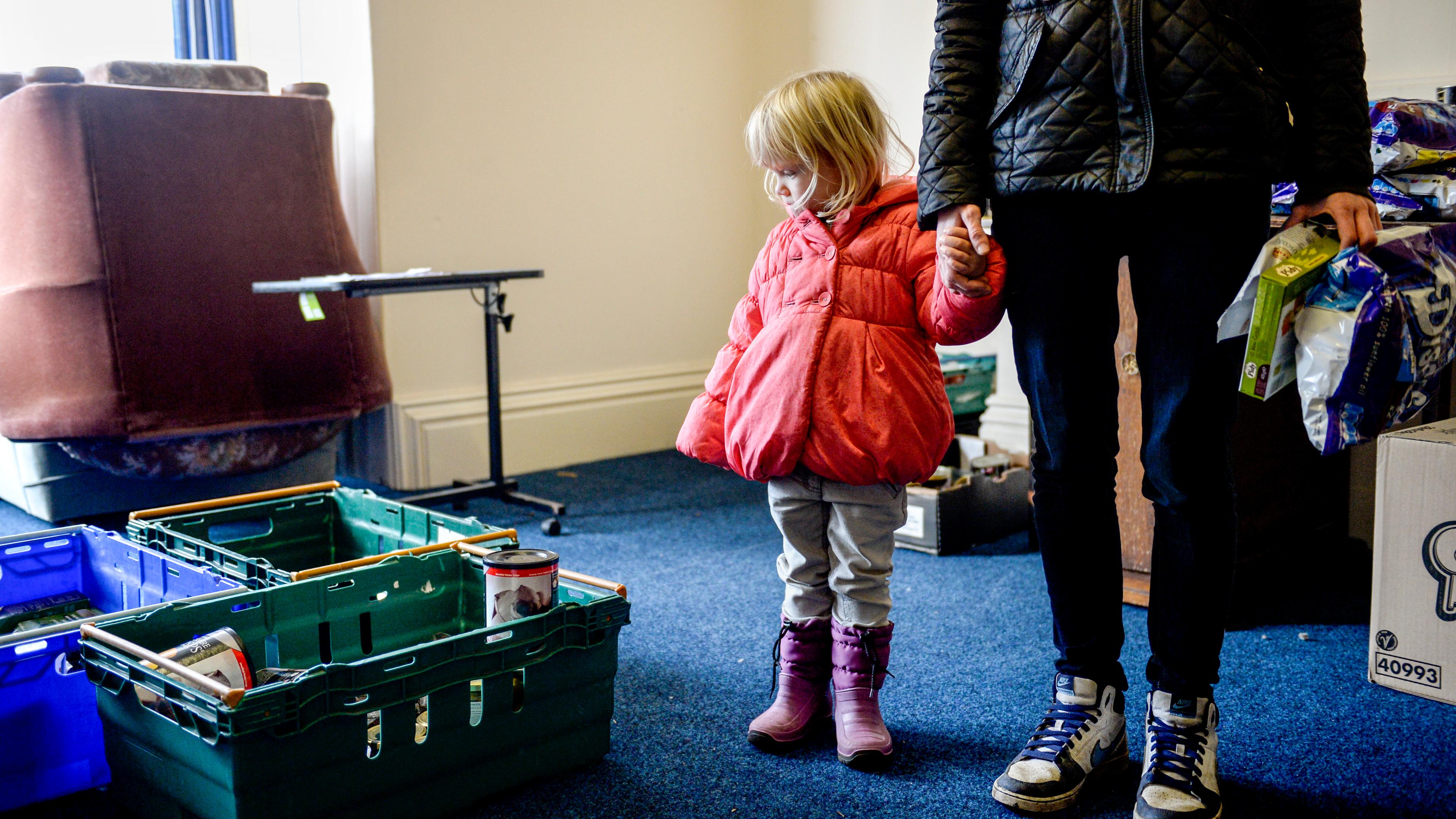 Photo of a child in a pink coat holding an adult’s hand in a room with crates containing canned goods.