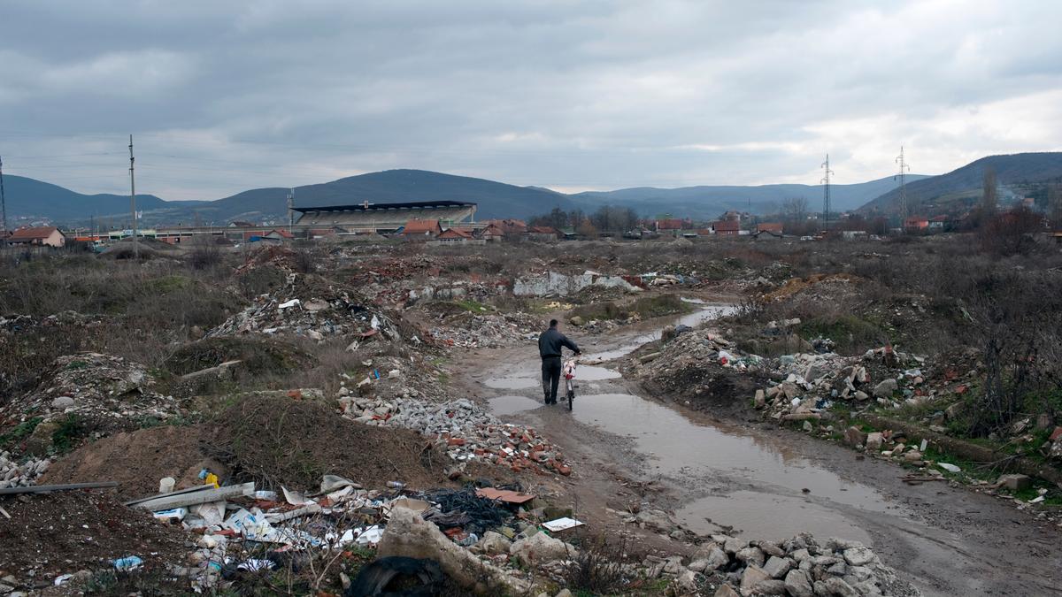 A person pushing a bike through a muddy landfill with debris under an overcast sky, distant hills and houses.