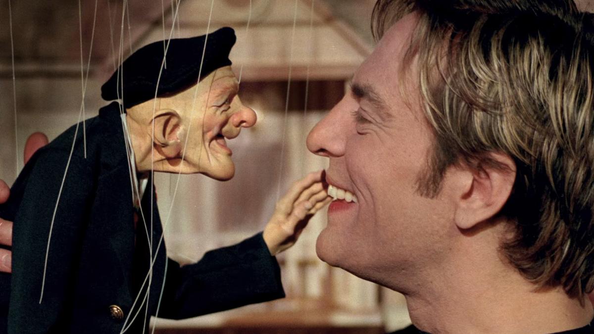 Close-up of a man smiling and interacting closely with a puppet of an old man wearing a black beret, both faces nearly touching.