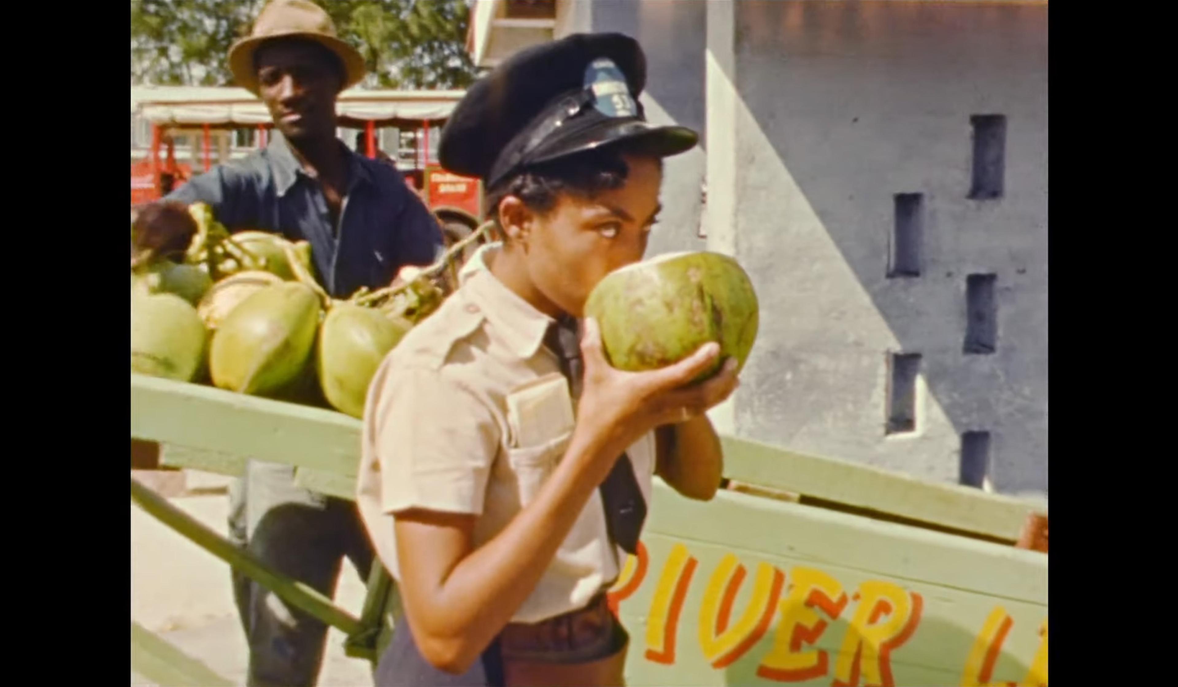 A female police officer drinks from a coconut while standing next to a vendor with a cart full of coconuts.