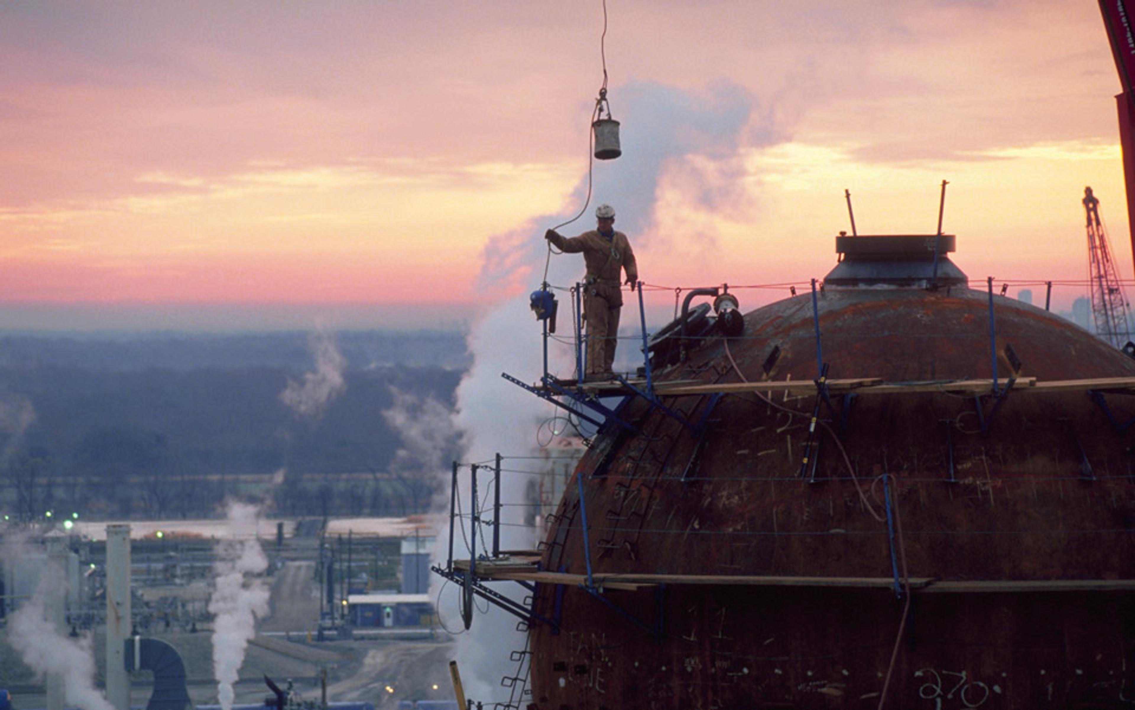 A worker standing on a large industrial sphere, with steam rising and a sunset in the background.
