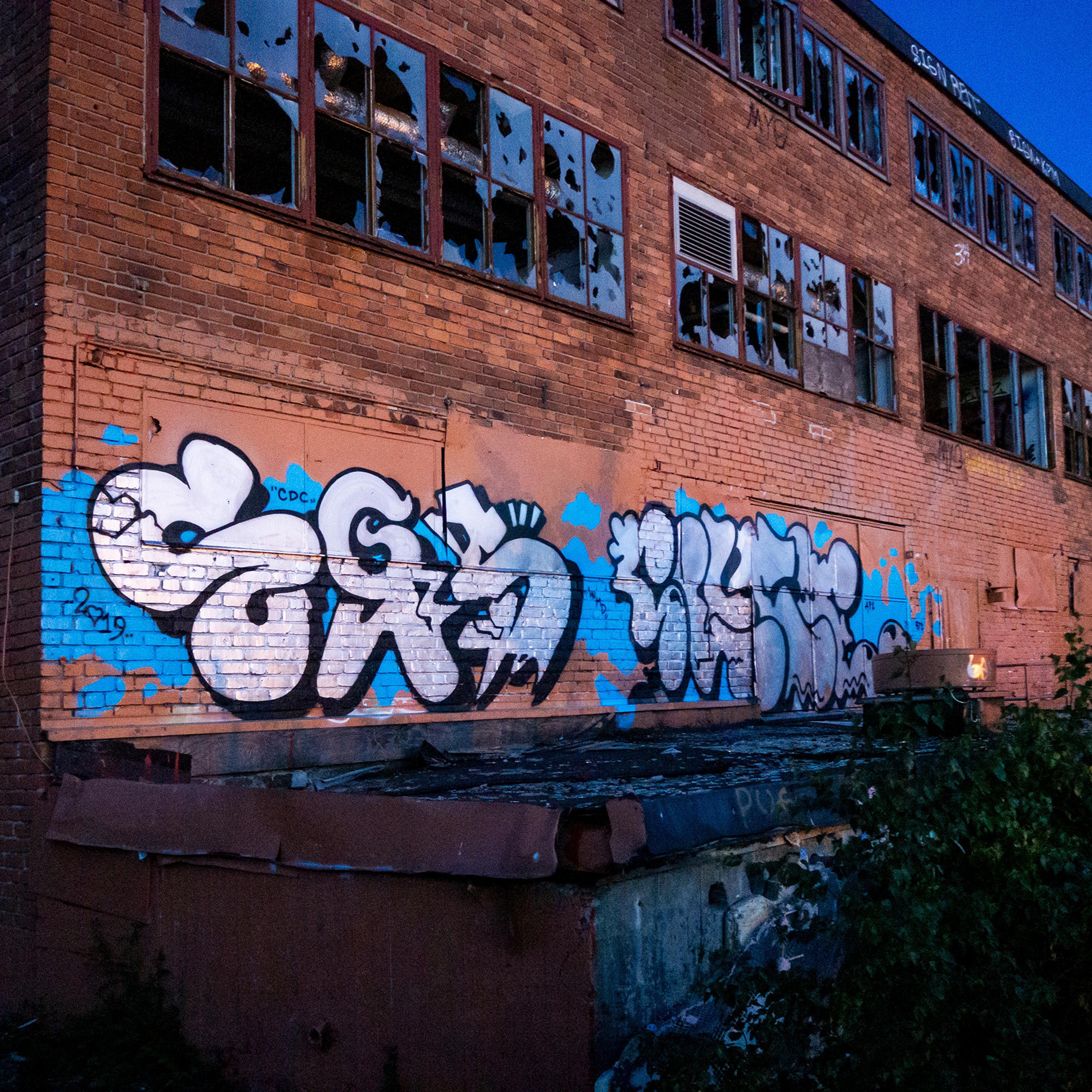 Photo of a derelict brick building with colourful graffiti on the wall broken windows and overgrown vegetation at dusk.