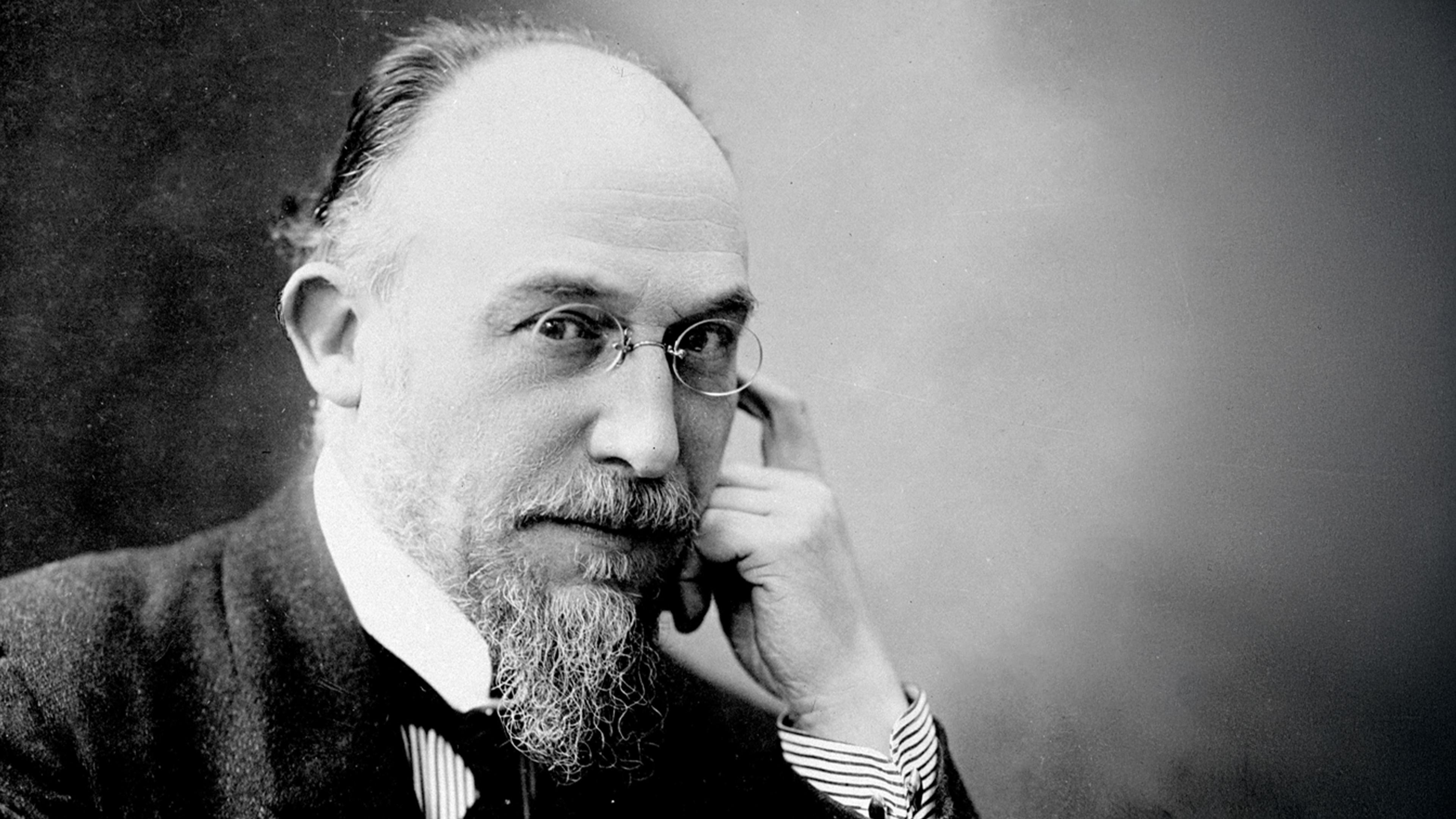 Black and white photo of a man with glasses and a beard, resting his chin on his hand, wearing a suit.