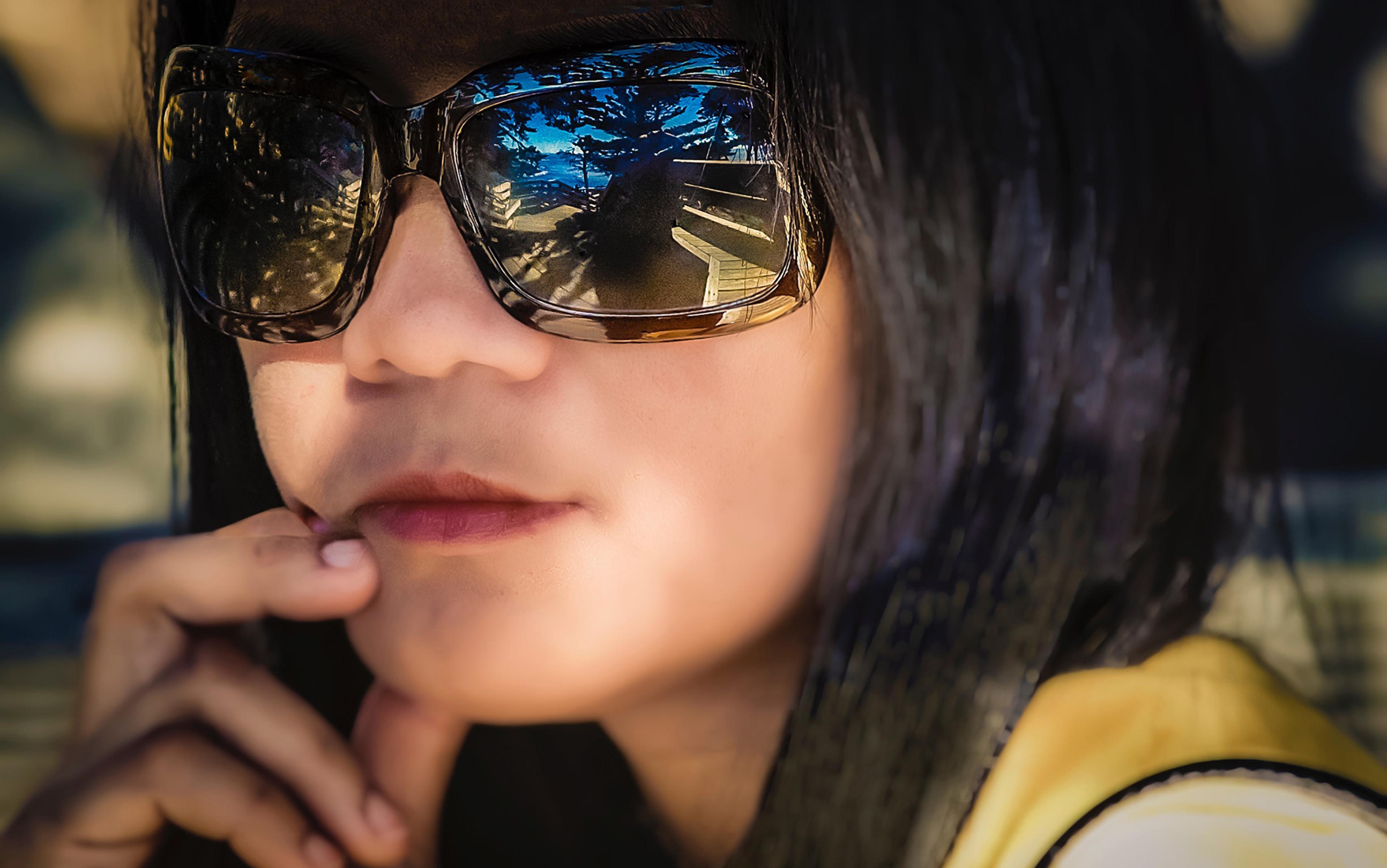 Photo of a person wearing large sunglasses with outdoor scenery reflected, resting their fingers on their chin in a thoughtful pose.