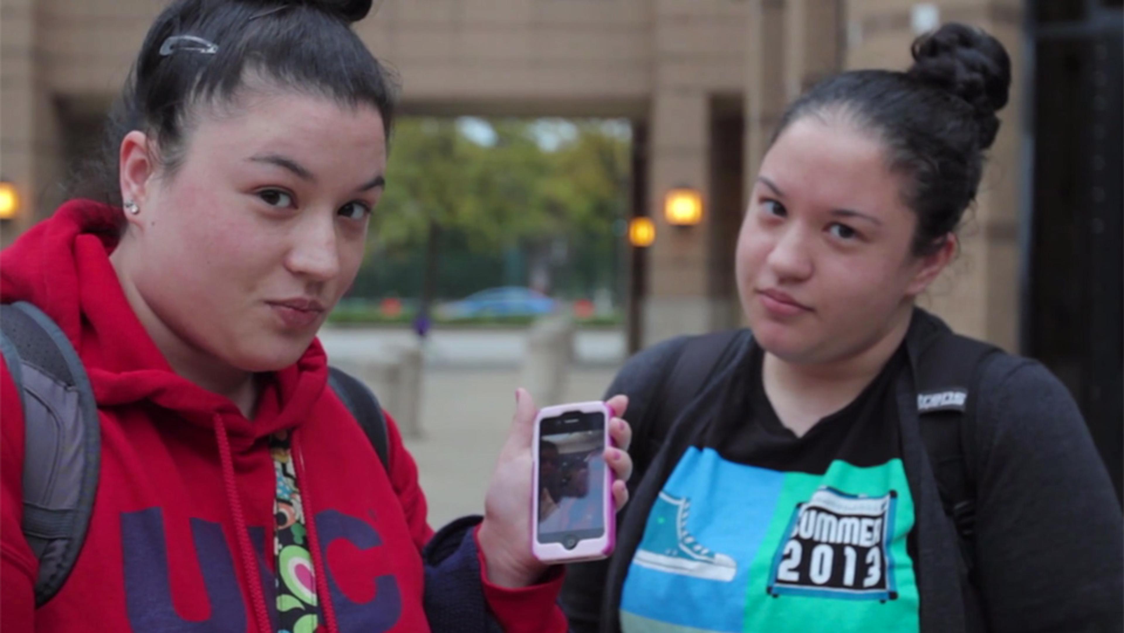 Two women with hair in buns, wearing backpacks, one in a red hoodie and the other in a black shirt, holding a mobile phone.