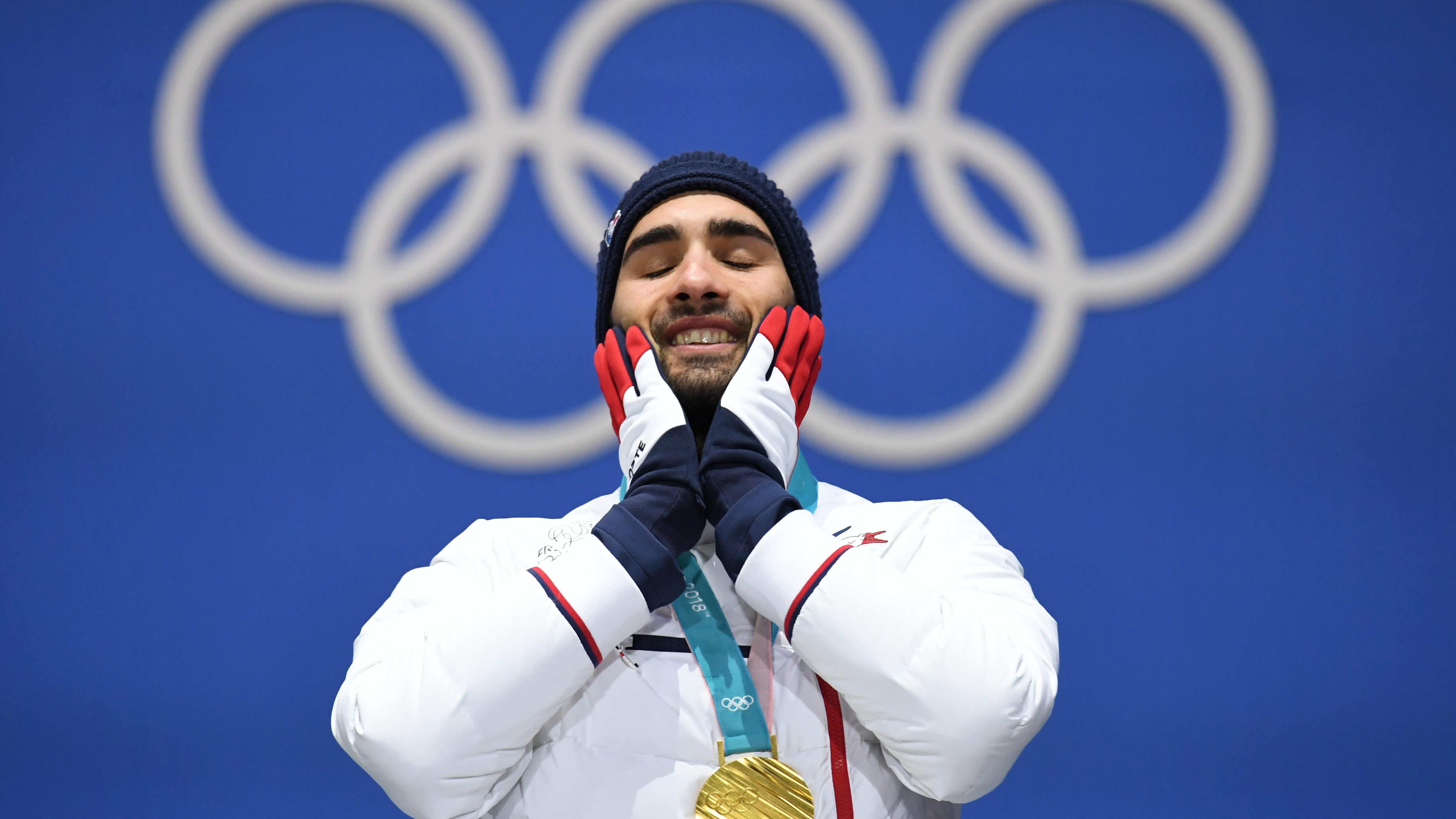 A joyful athlete wearing a gold medal holds his hands to his cheeks standing in front of an Olympic rings background.