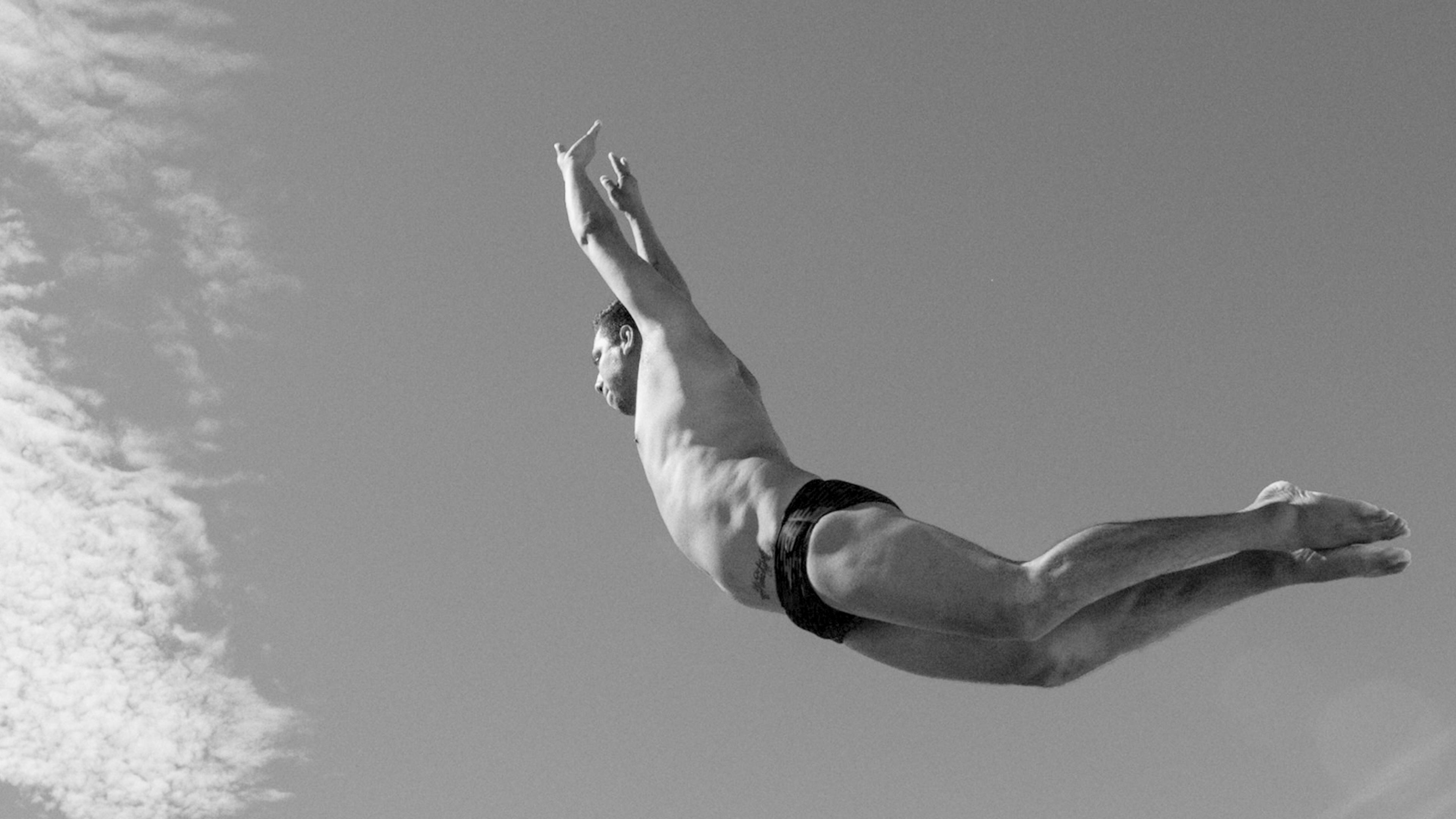 Black-and-white photo of a man diving mid-air against the sky, arms extended and wearing swim briefs.