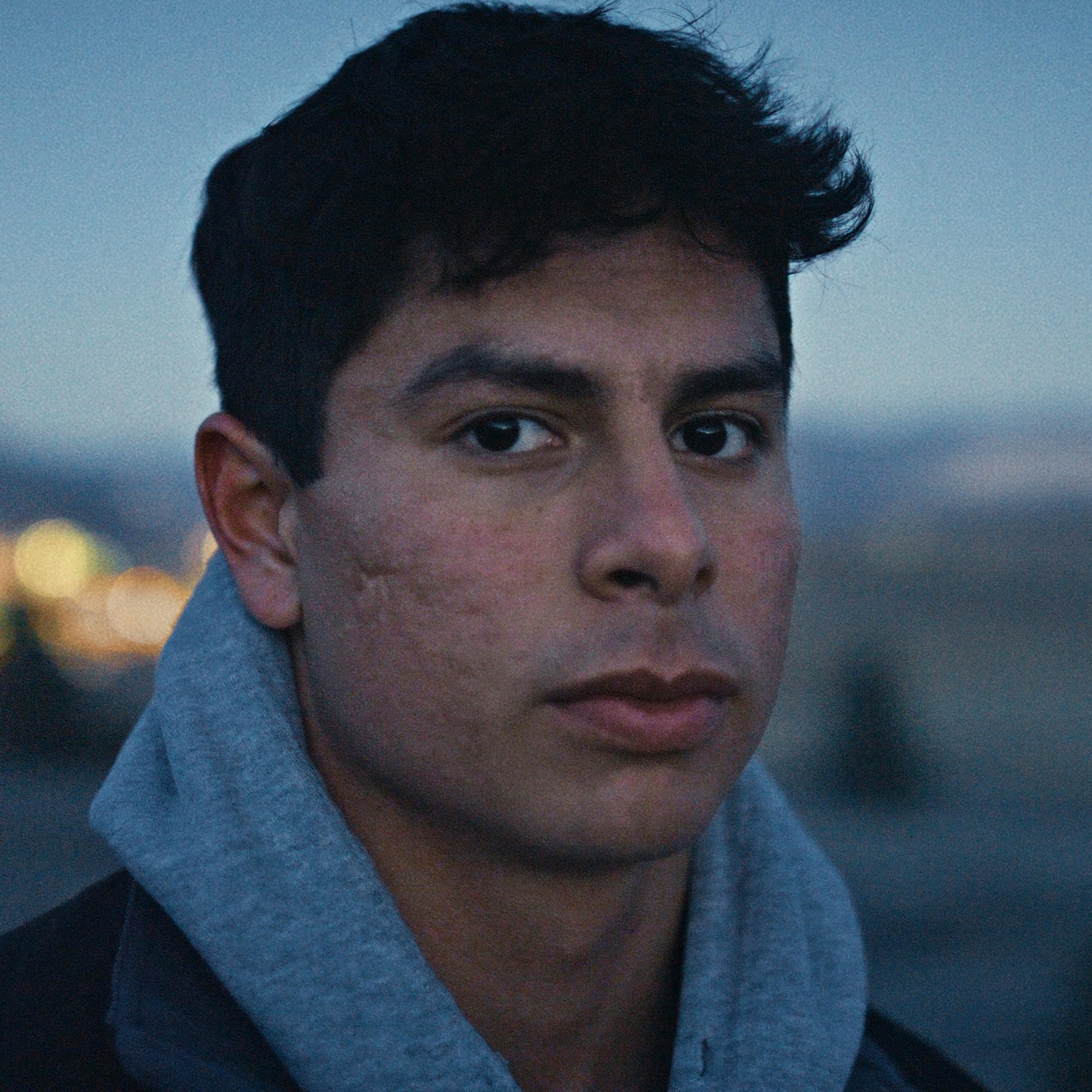 A young man with a serious expression wearing a grey hoodie outdoors at dusk, with a blurred landscape and lights in the background.