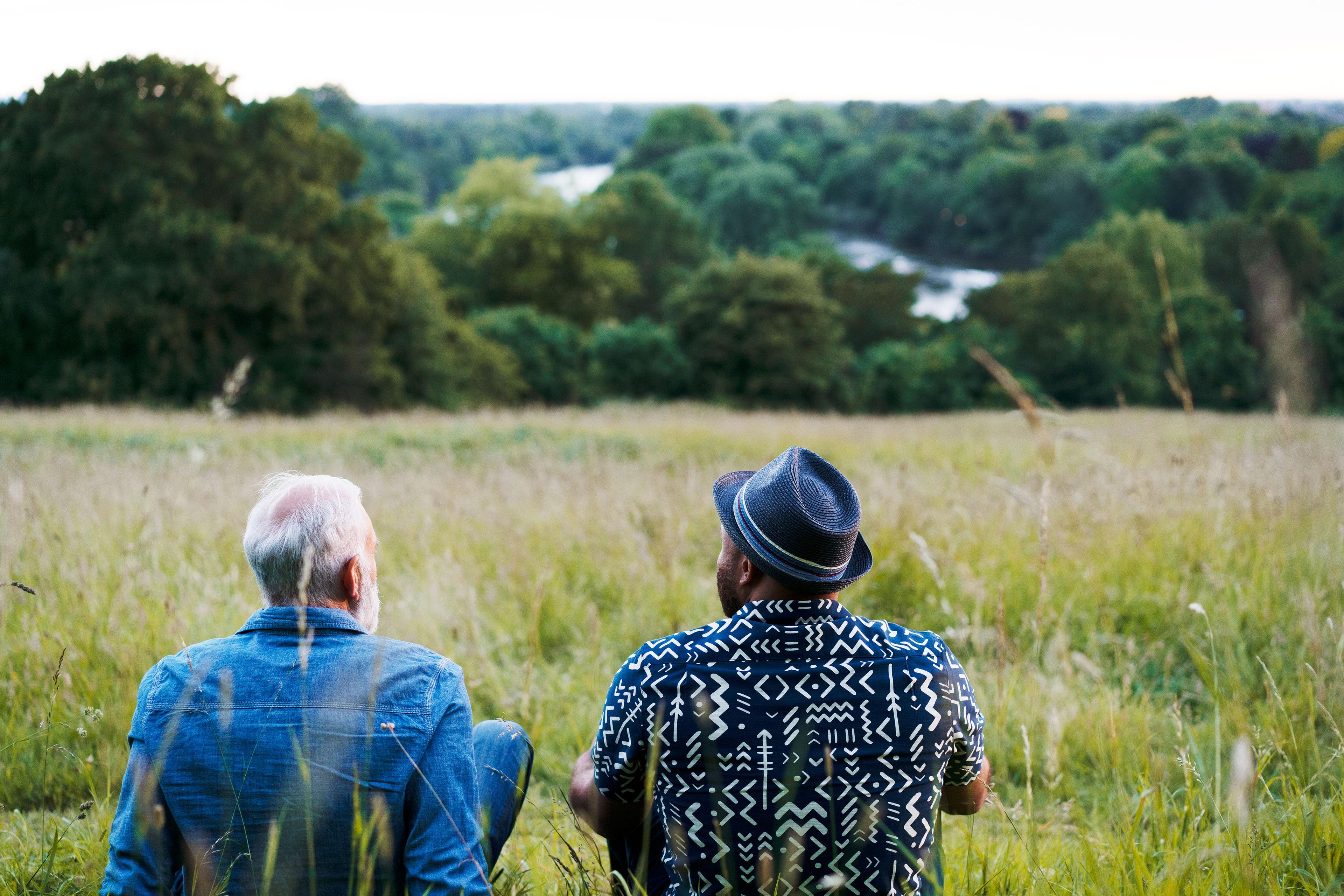 Photo of two people sitting in a field overlooking a river and trees with one wearing a hat and the other in a denim jacket.