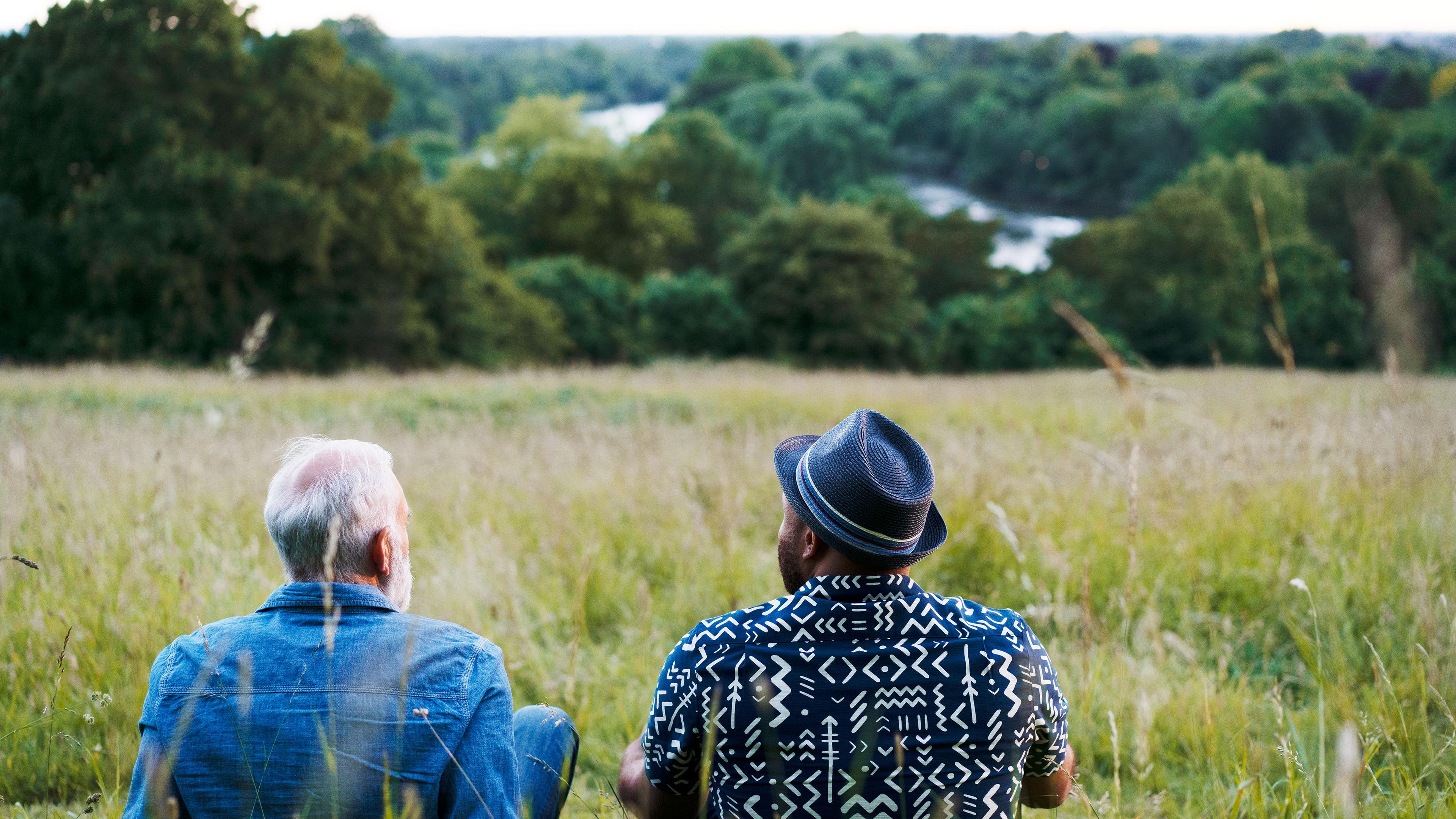 Photo of two people sitting in a field overlooking a river and trees with one wearing a hat and the other in a denim jacket.