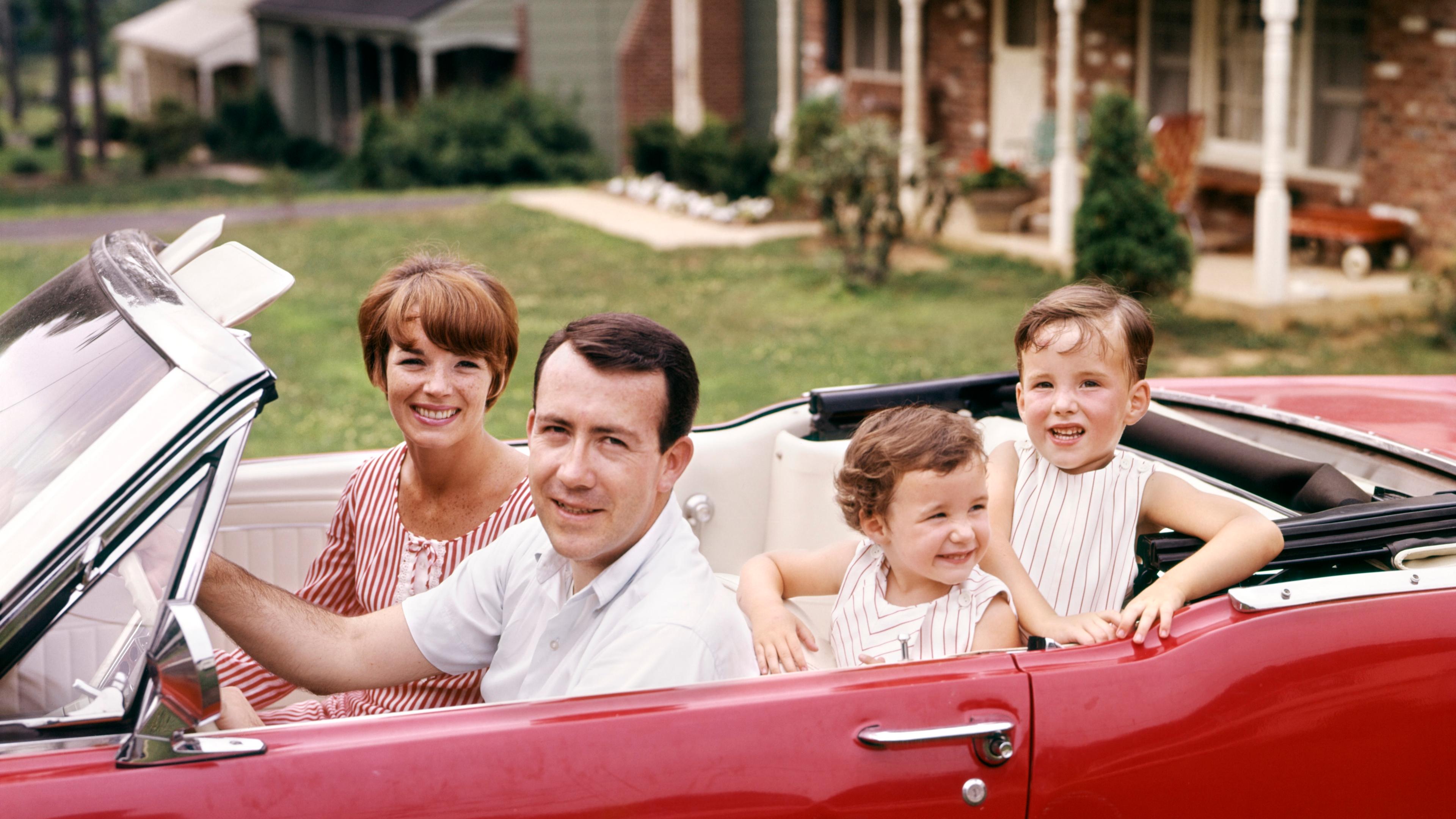 Photo of a smiling family sitting in a red convertible car parked in front of a suburban house.