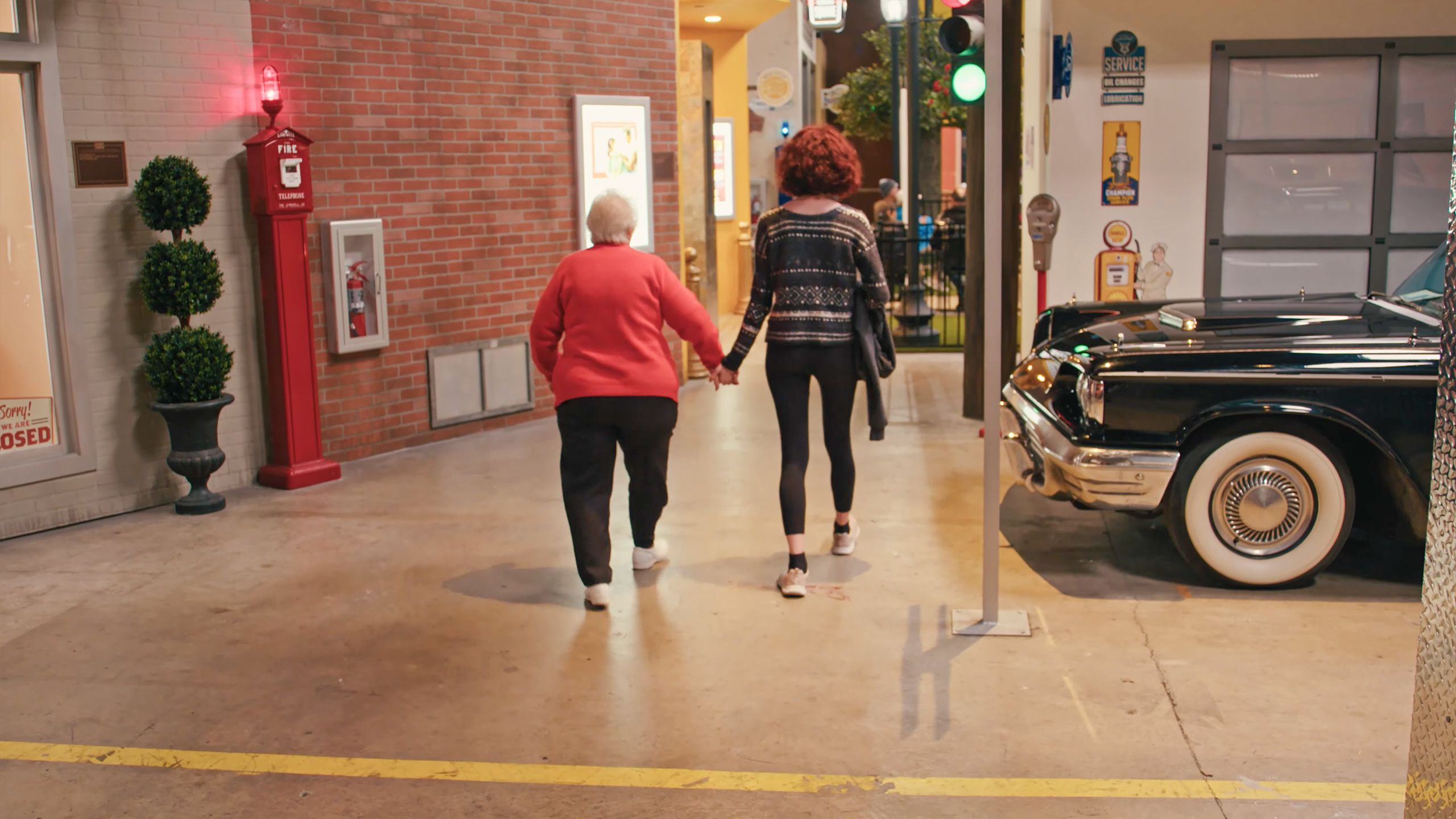Photo of an elderly woman and a teenager walking hand in hand inside a building with retro decor, a vintage car beside them.