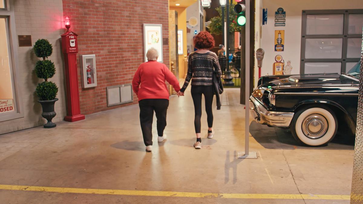 Photo of an elderly woman and a teenager walking hand in hand inside a building with retro decor, a vintage car beside them.