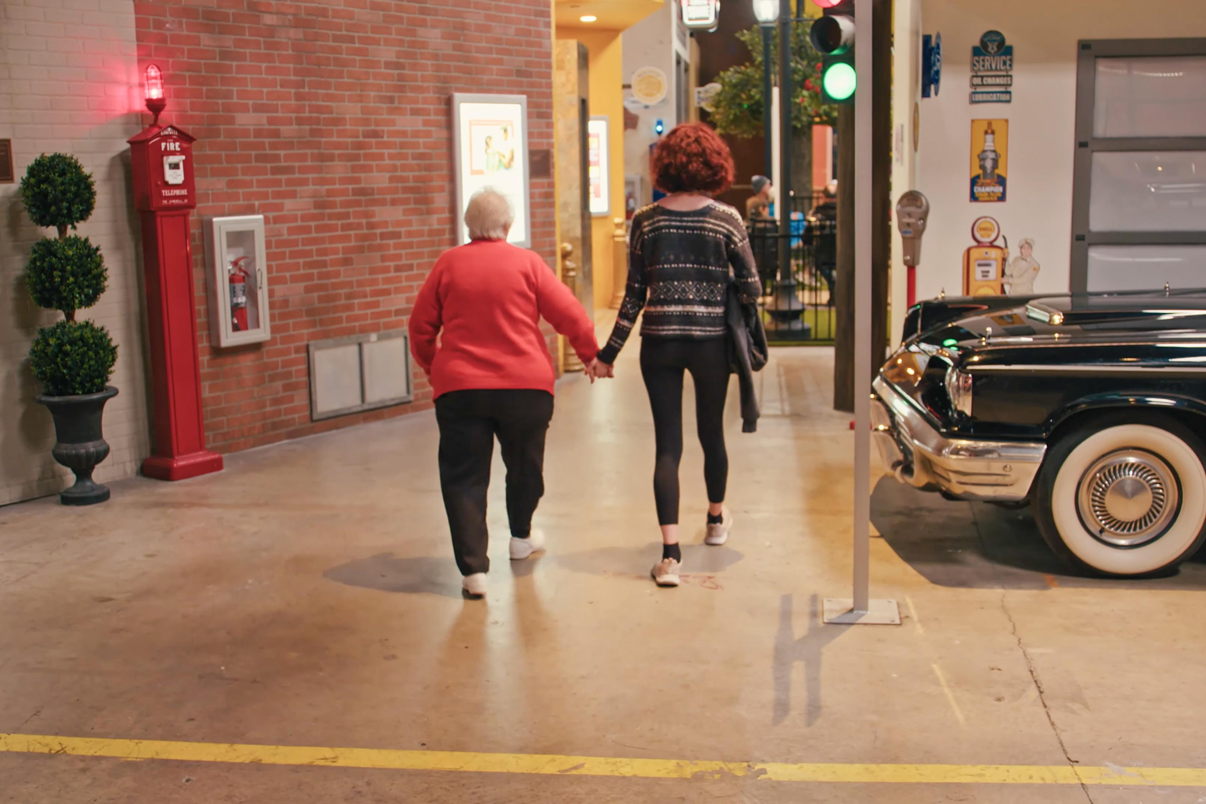 Photo of an elderly woman and a teenager walking hand in hand inside a building with retro decor, a vintage car beside them.