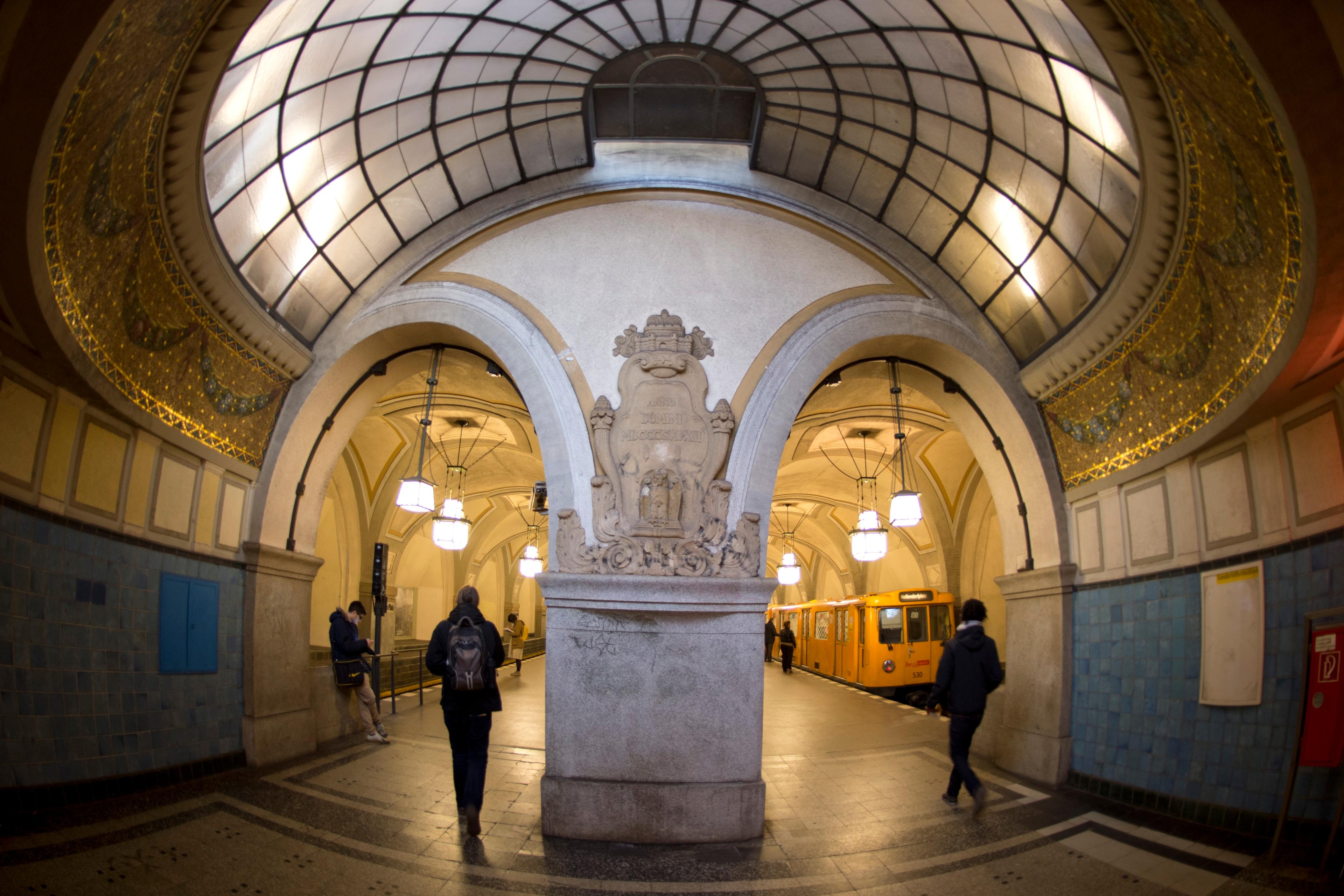 Photo of an ornate underground station with arched ceilings, mosaic details pedestrians and a yellow train on the platform.