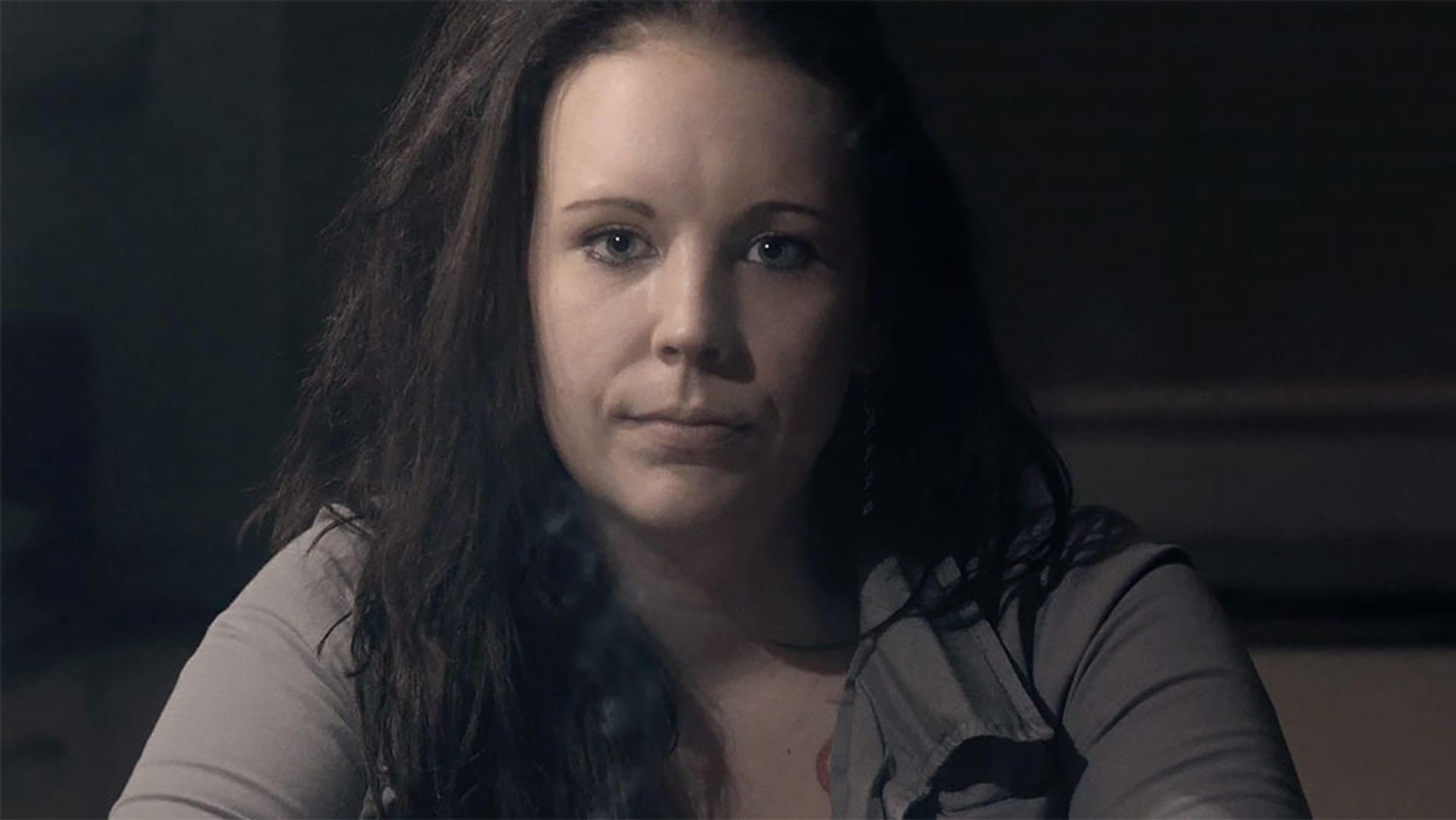 A woman with long hair wearing a grey shirt, looking directly at the camera, seated indoors with a dark background.