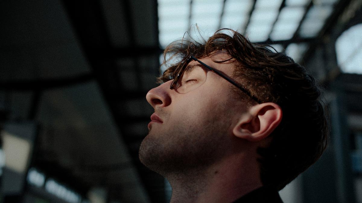 Photo of a man in profile with glasses, eyes closed, head tilted back under a structure with a grid-like roof.