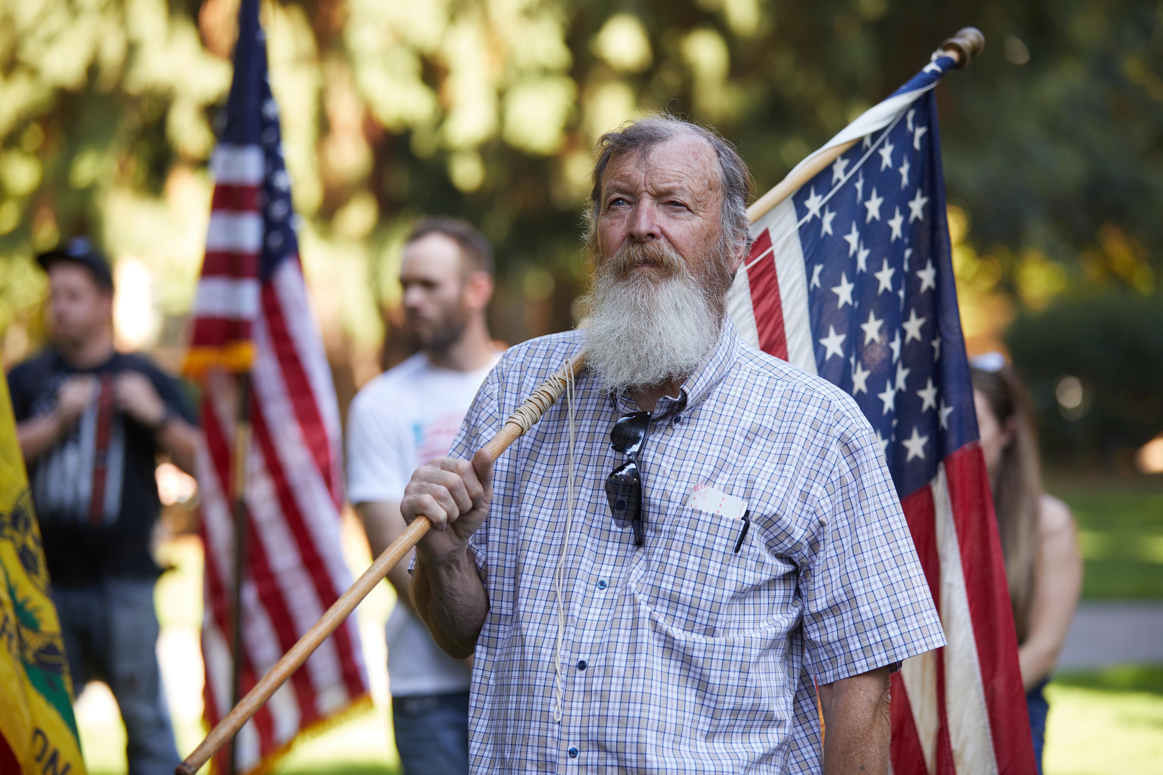 Photo of a bearded man holding a US flag at an outdoor gathering, with blurred people and flags in the background.