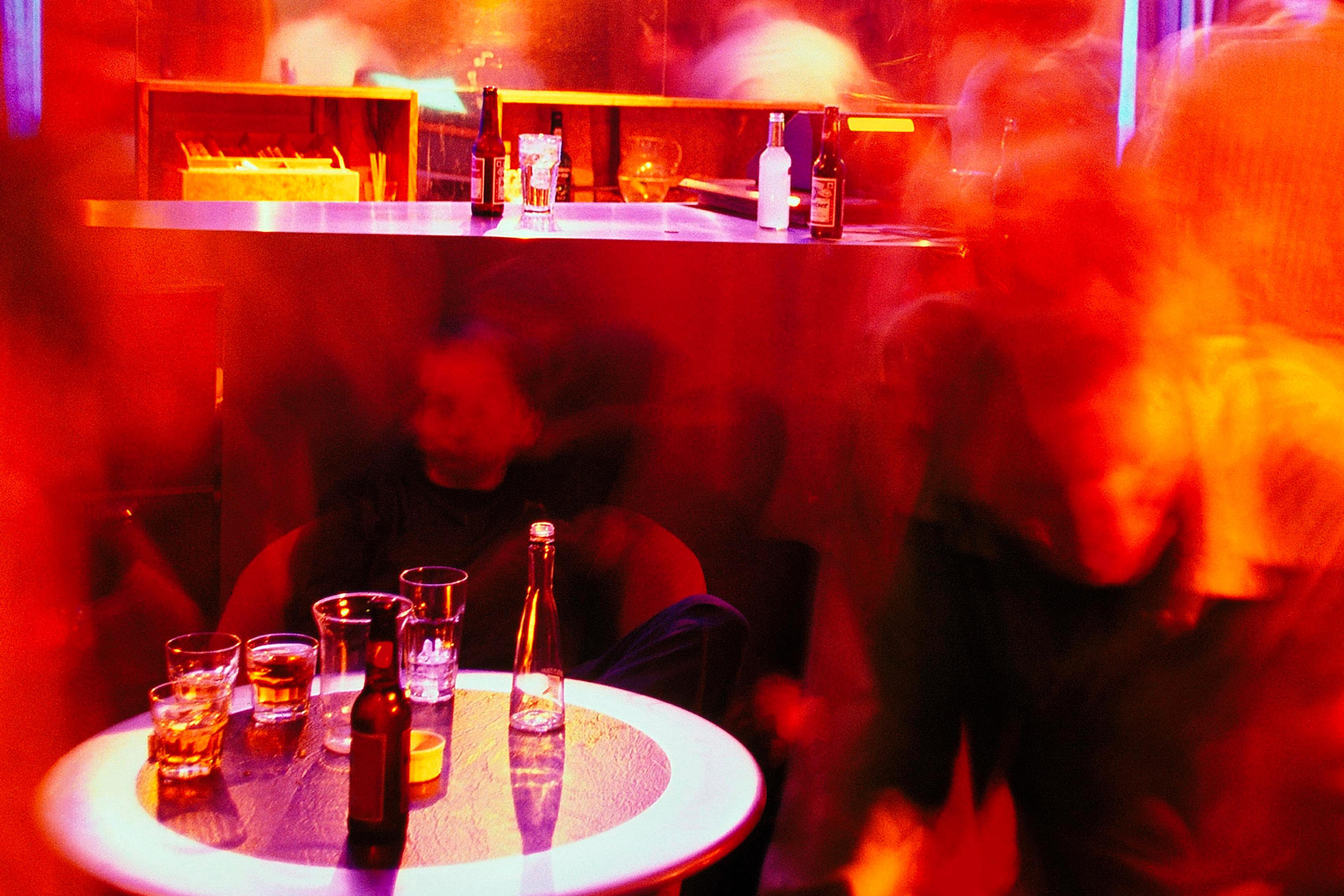 Blurry photo of a vibrant bar scene with a table of drinks, red lighting and indistinct figures moving around.