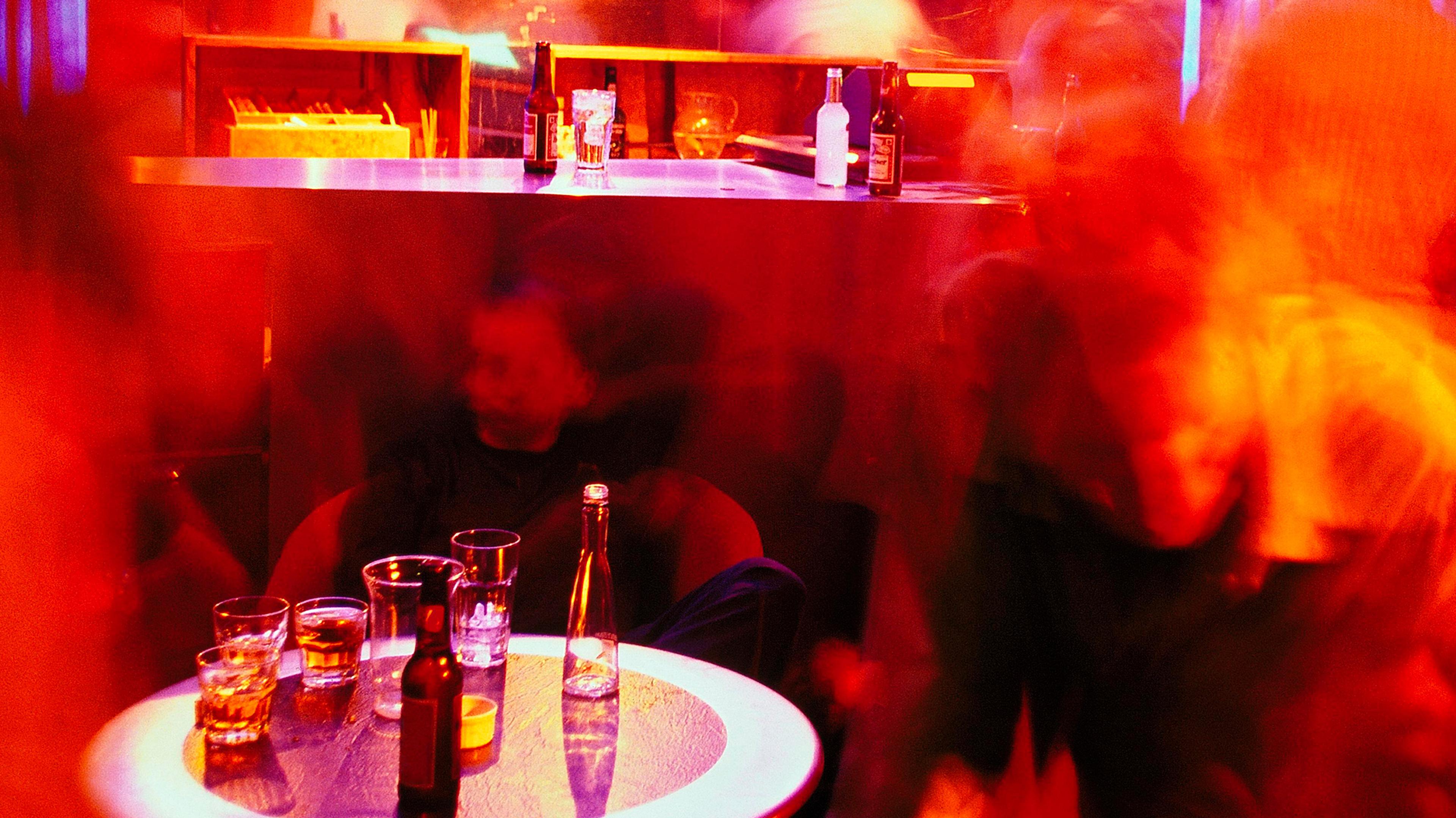 Blurry photo of a vibrant bar scene with a table of drinks, red lighting and indistinct figures moving around.