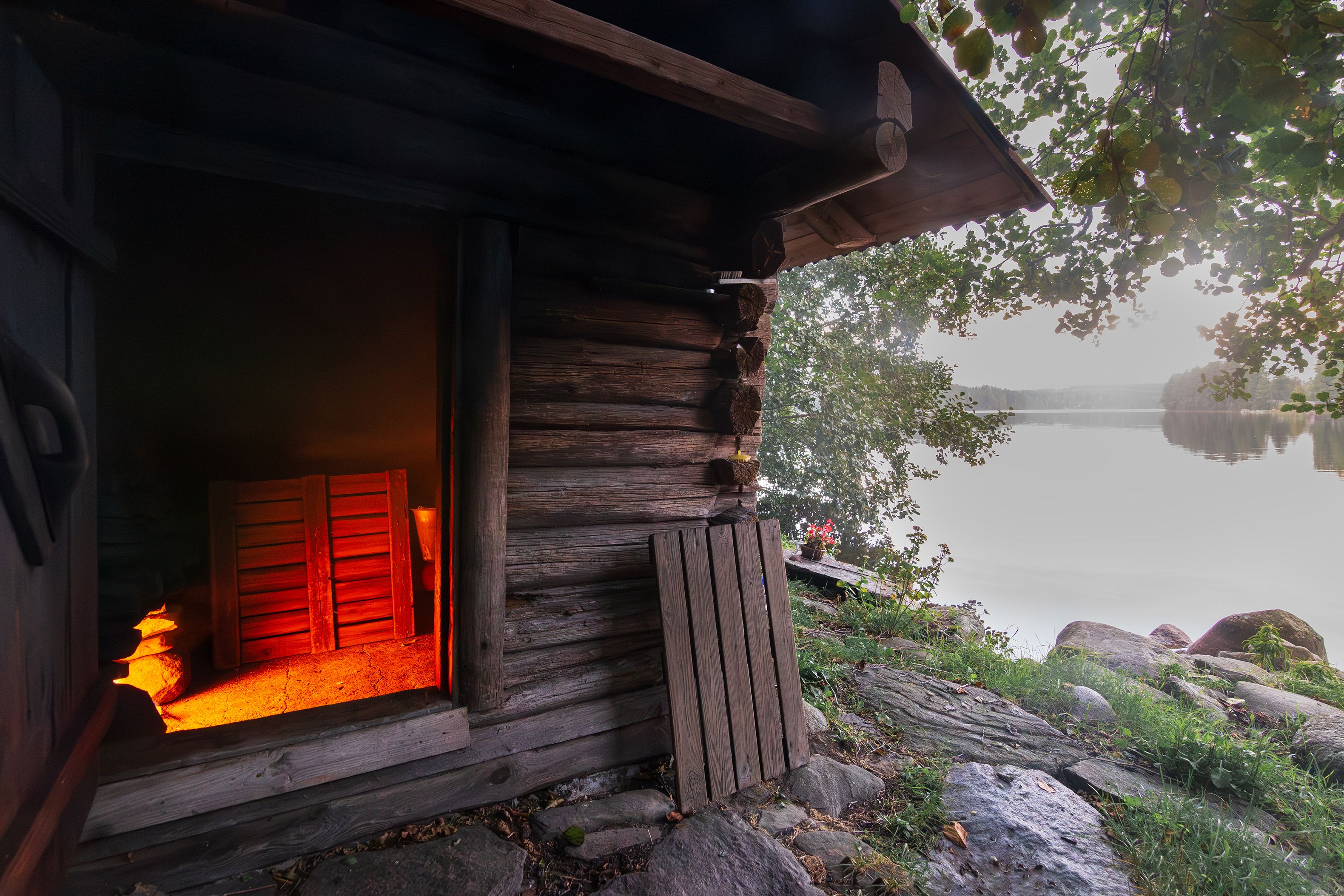 A rustic log sauna by a lake. Warm orange glow from inside contrasts with the serene lakeside view.