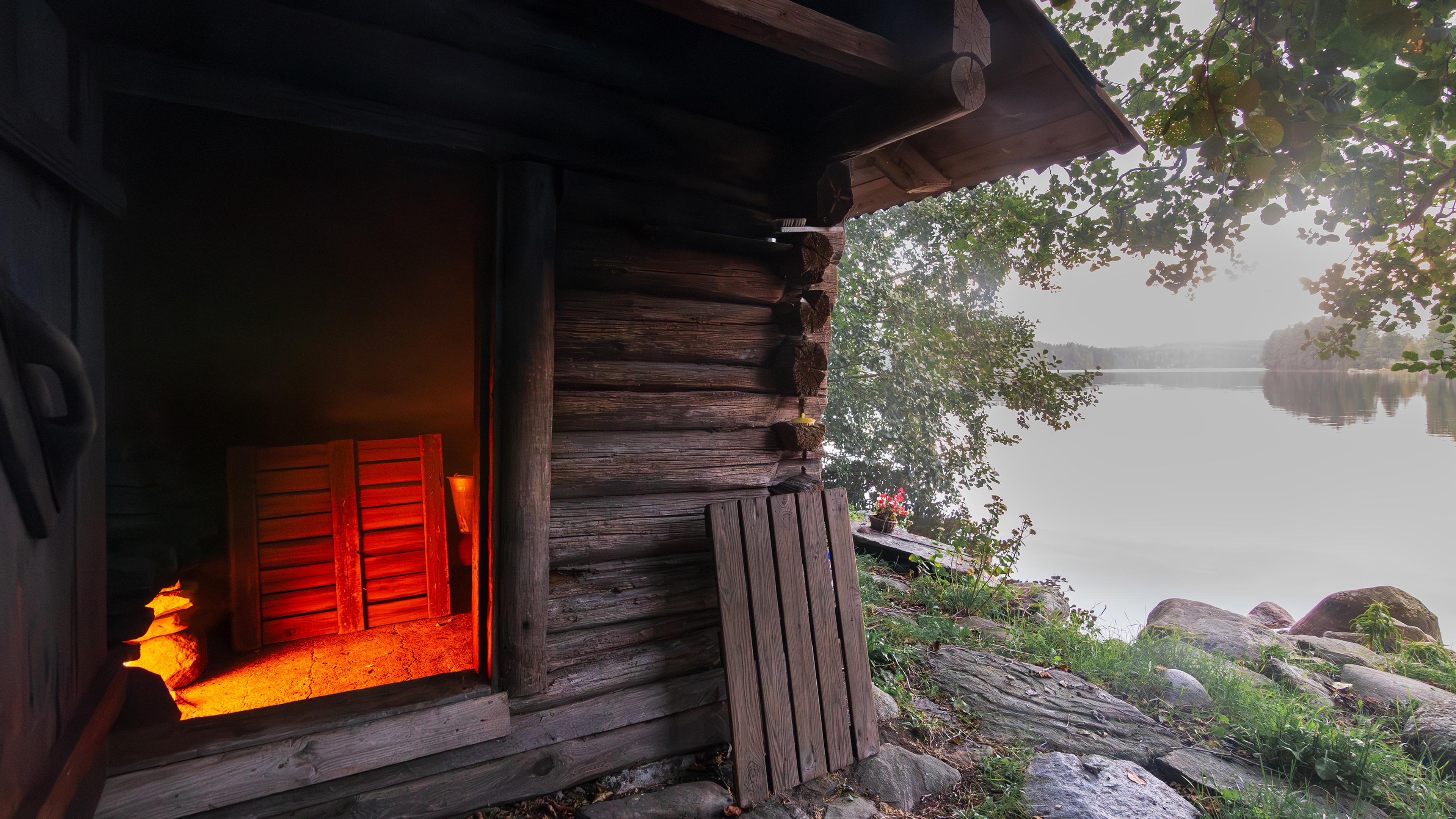 A rustic log sauna by a lake. Warm orange glow from inside contrasts with the serene lakeside view.