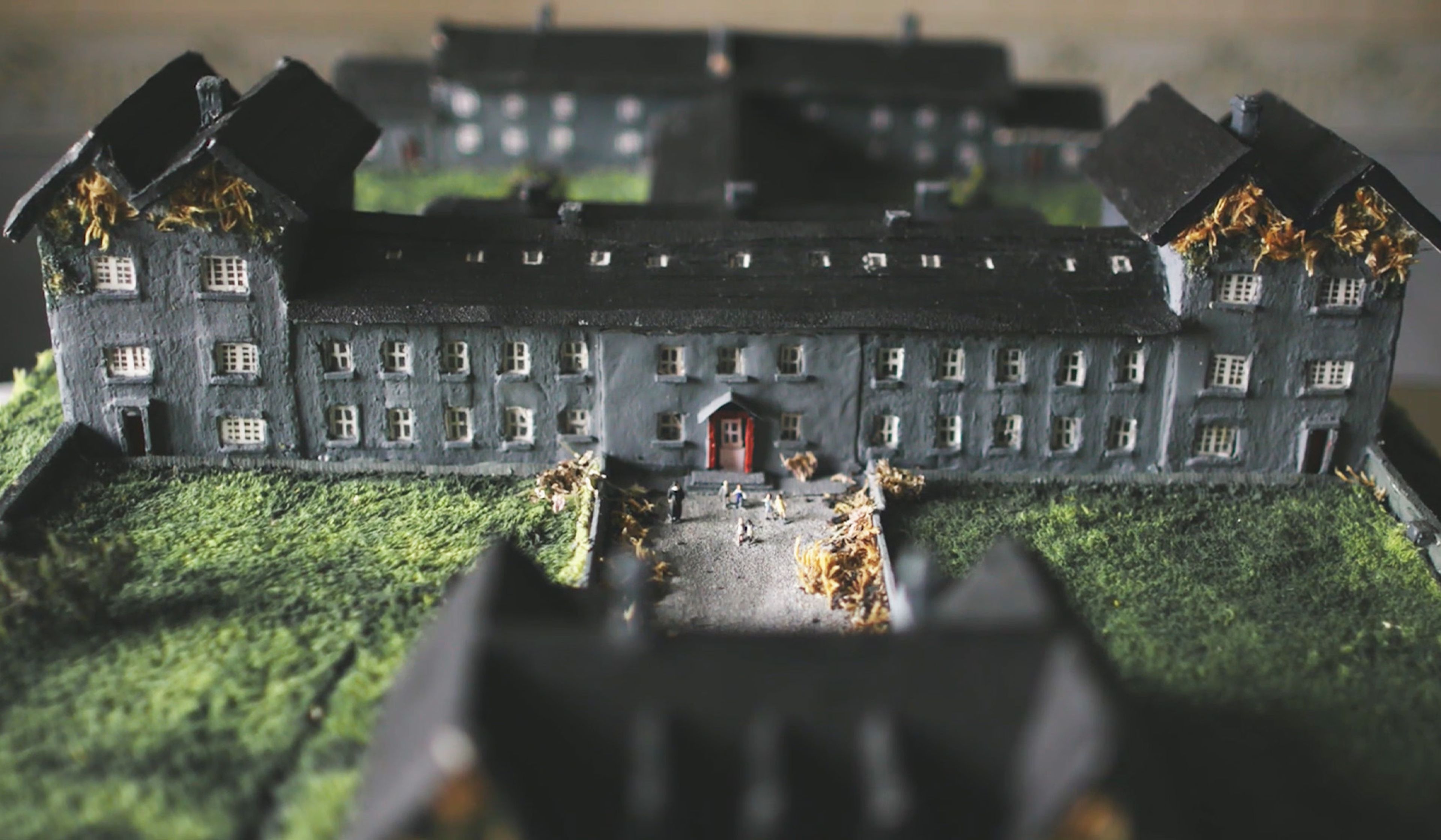 Model of a grey building with a red door, surrounded by grass and small figurines, representing an institution.