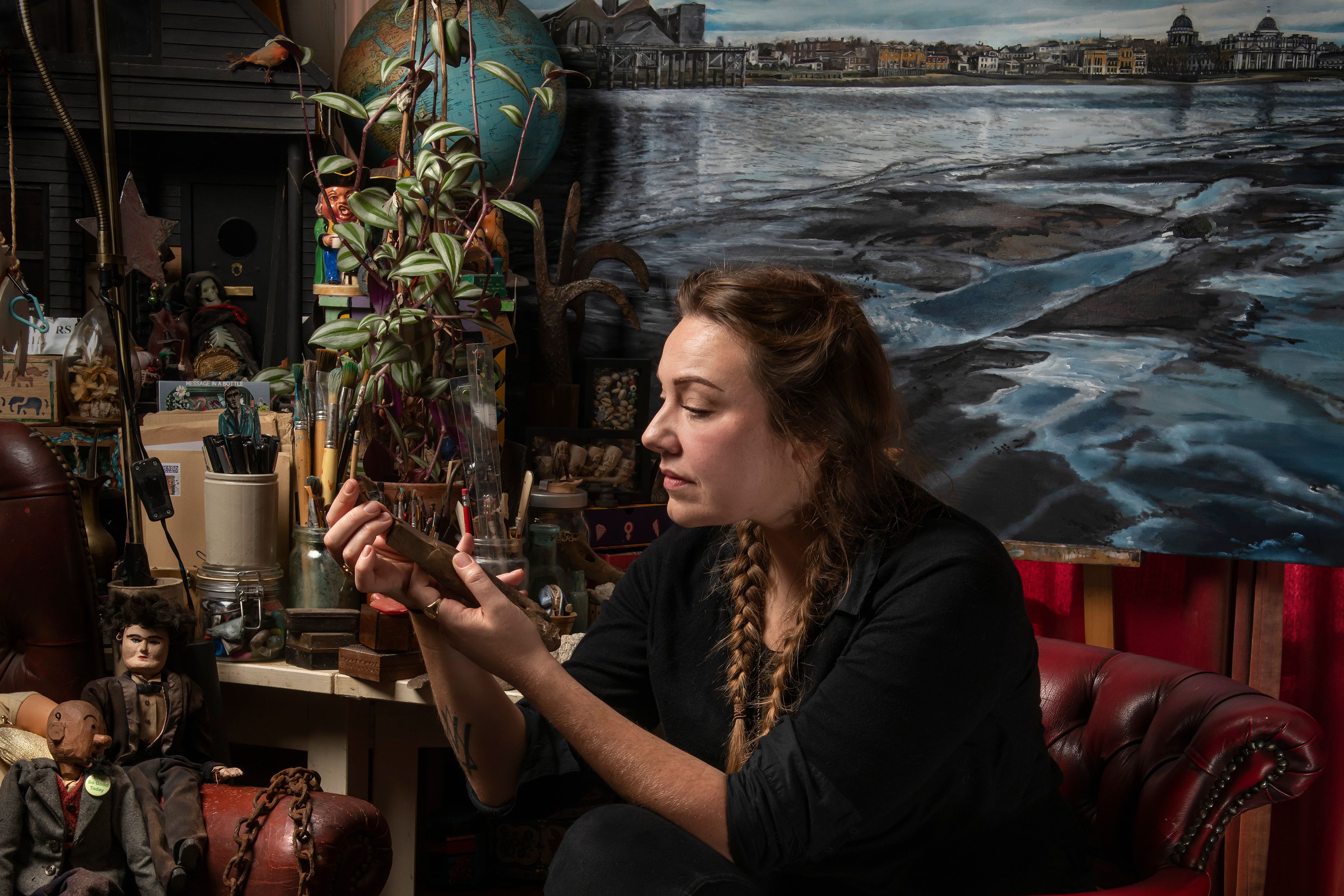 A woman in an art studio examining a small object surrounded by paintings, plants and art supplies.