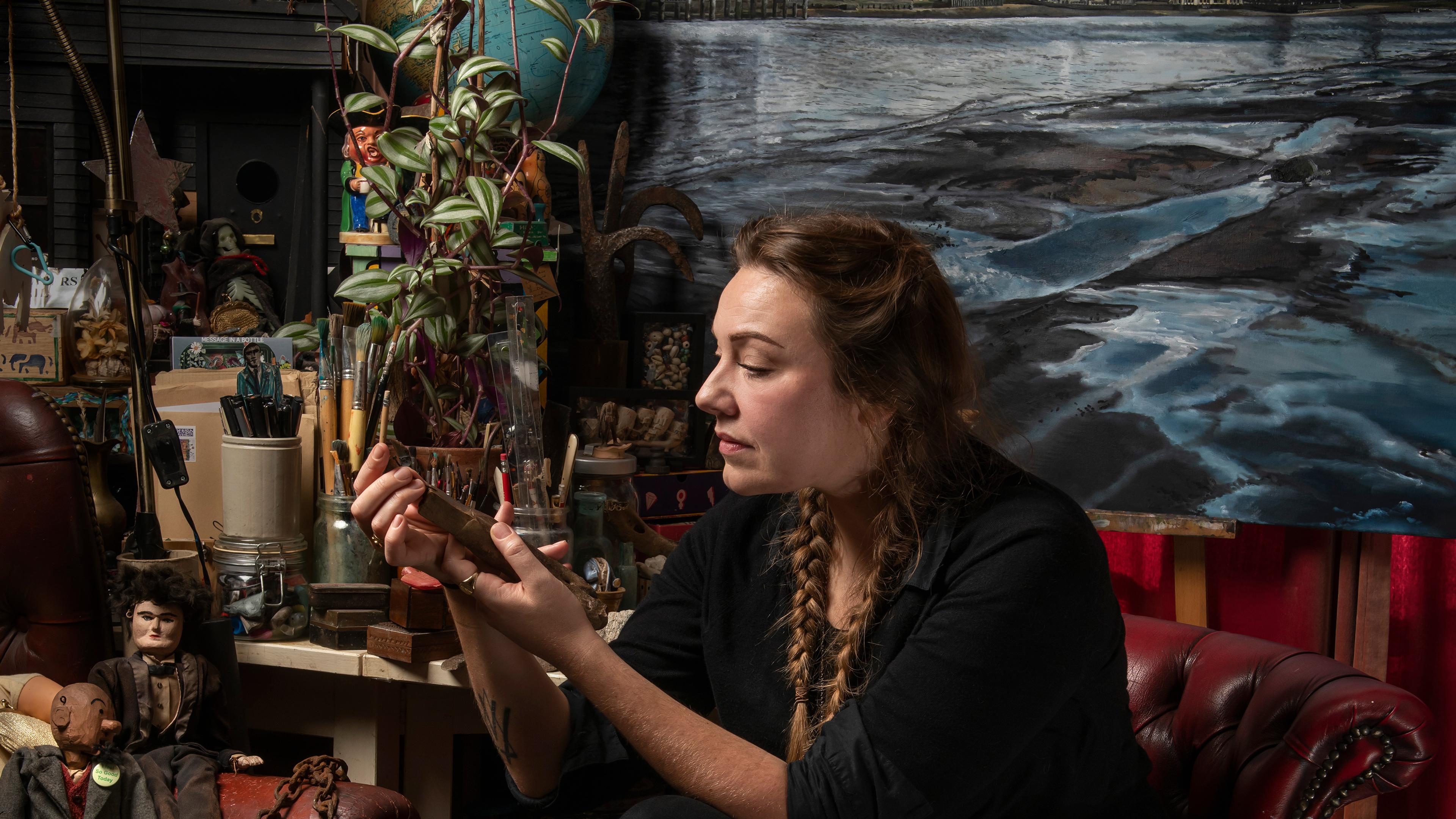 A woman in an art studio examining a small object surrounded by paintings, plants and art supplies.
