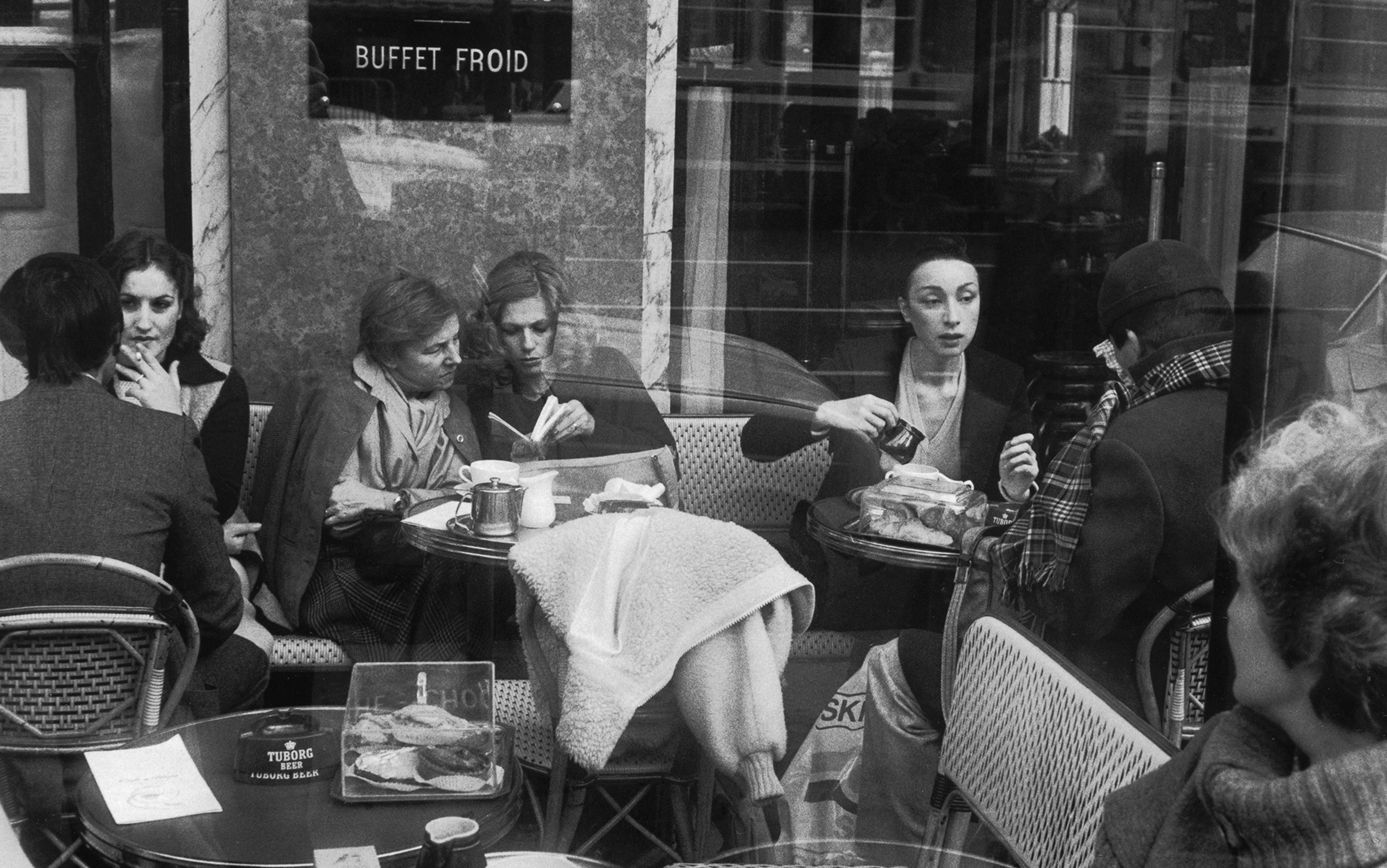 Black and white photo of people sitting at a café, taken through a window with reflections. A sign saying ‘BUFFET FROID’ is visible.
