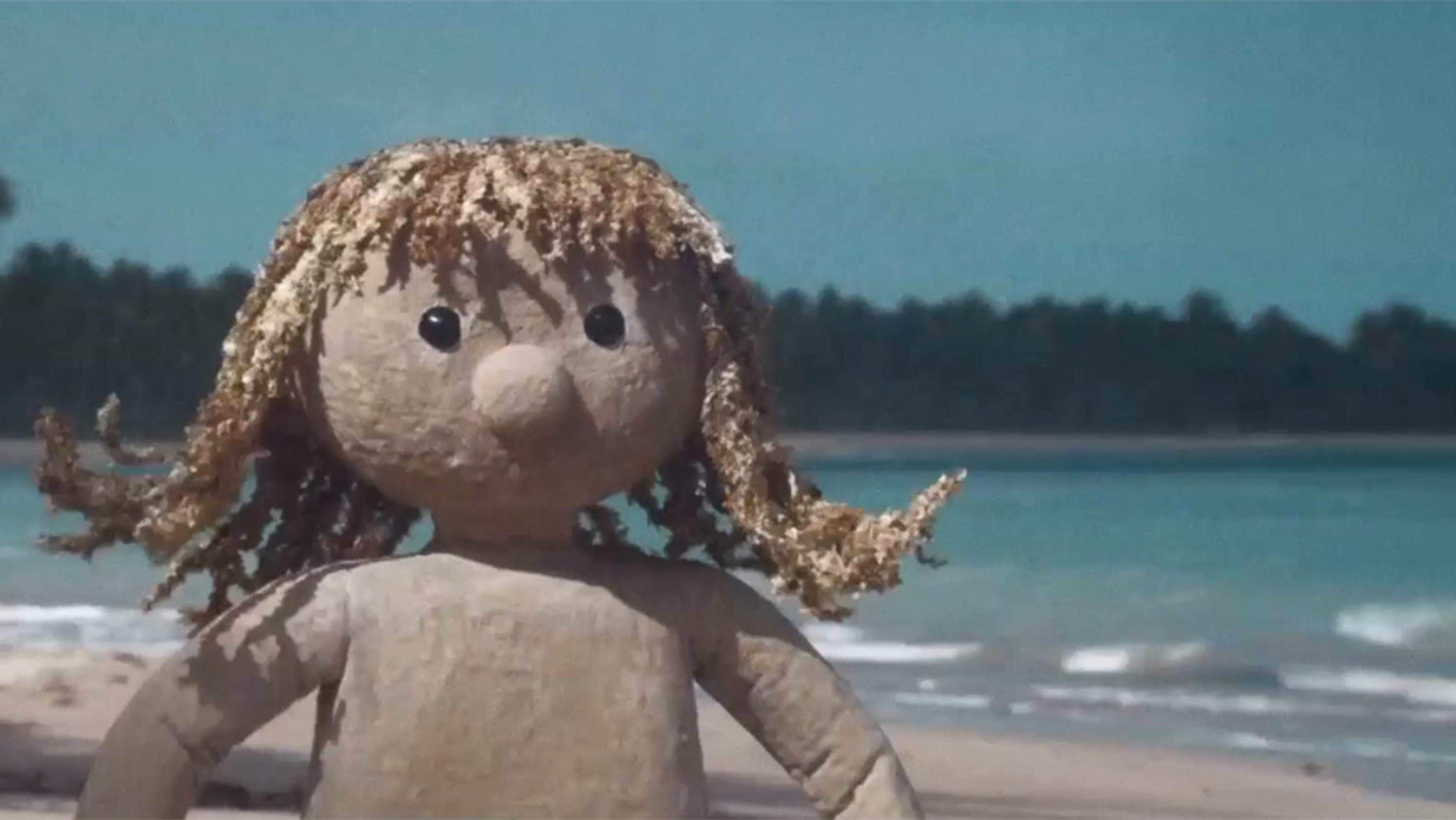 A simple puppet with light curly hair and black shiny eyes stands on a beach in front of the sea, with trees visible on the shoreline.