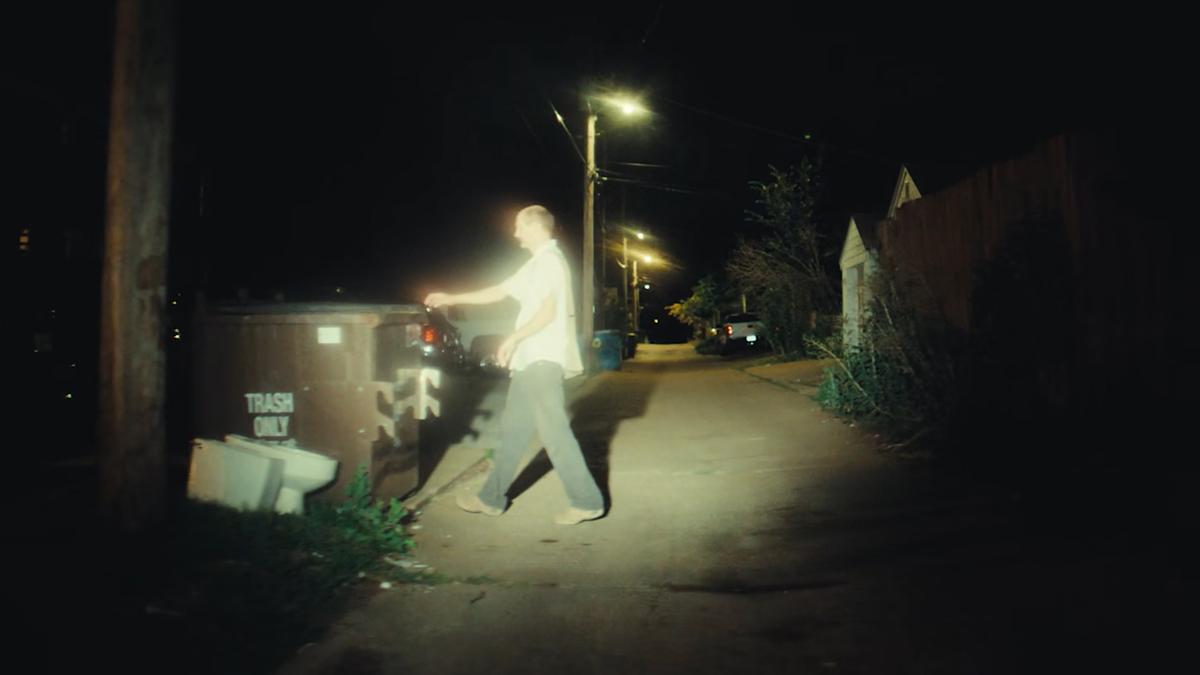 A person at night placing an object in a large bin labelled ‘TRASH ONLY’ on an empty street lit by streetlights.