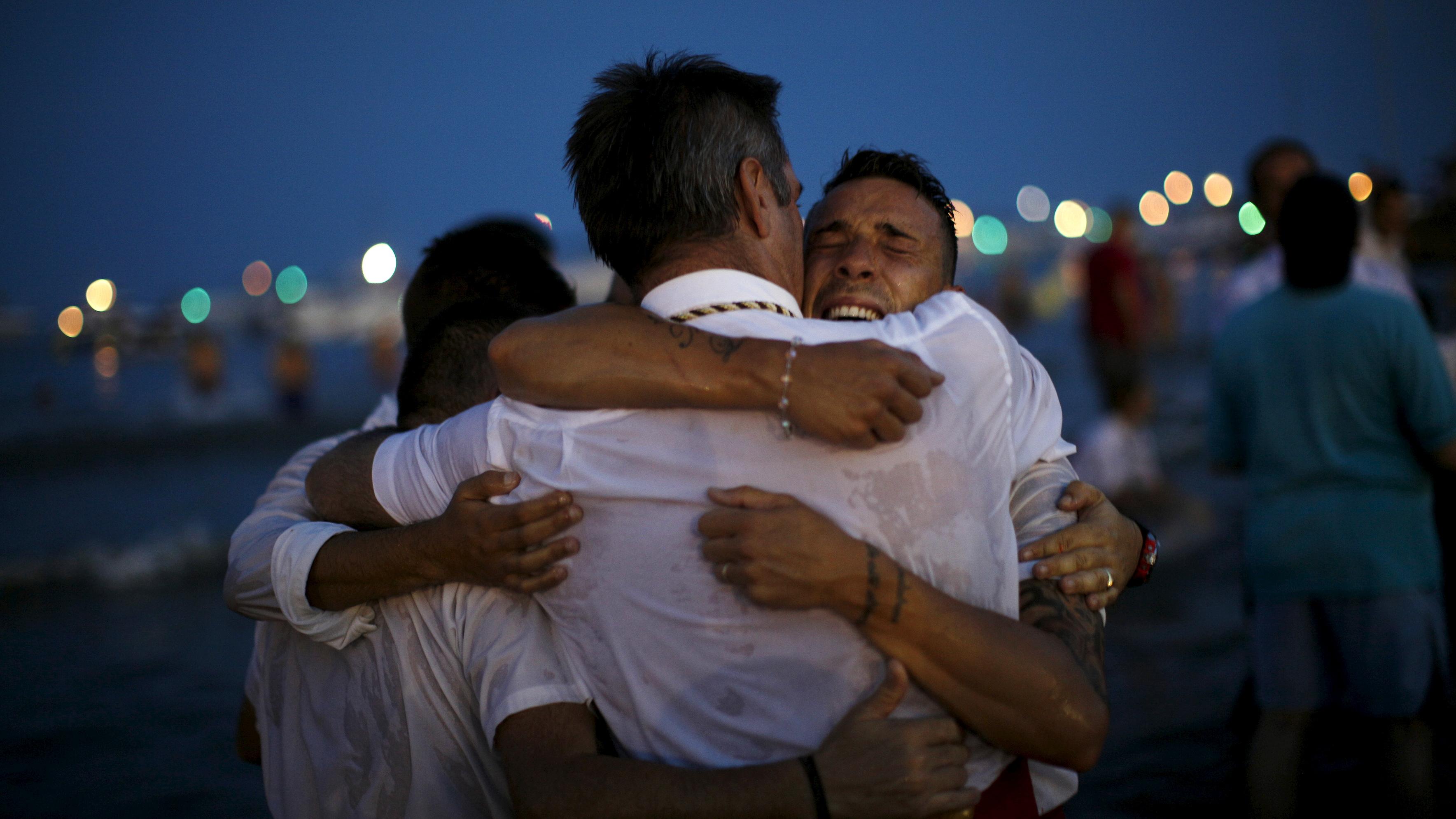 Photo of four men embracing on a beach at night with colourful lights blurred in the background.