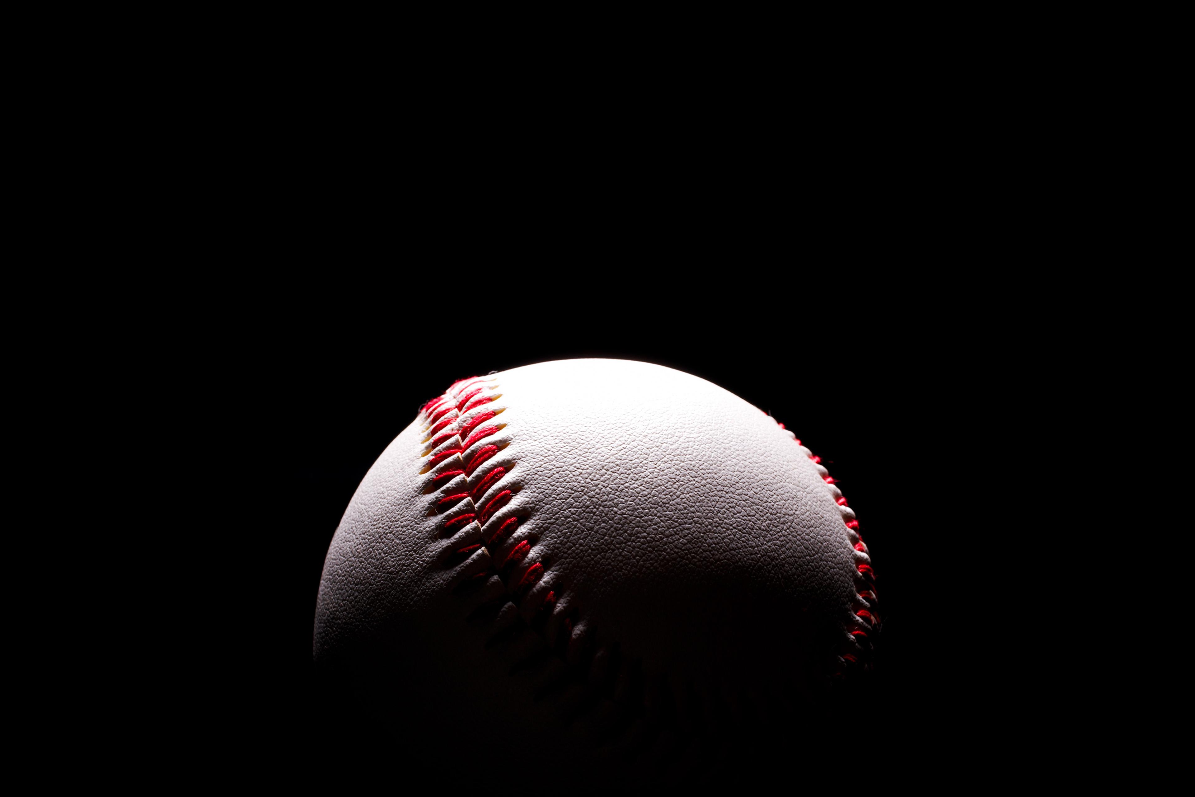 A baseball with red stitching on a black background partially lit from the top left corner.