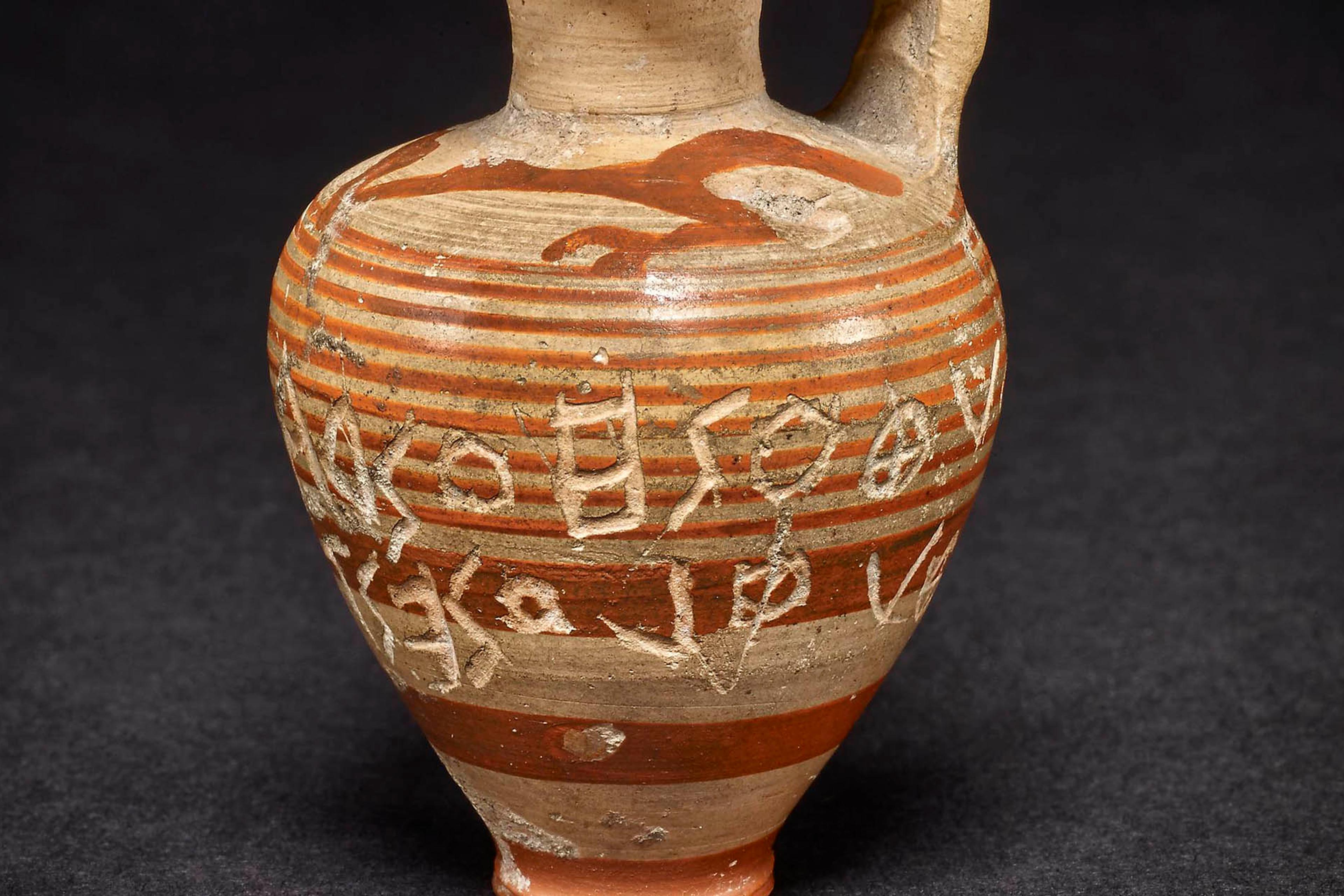 An ancient Greek clay jug with red stripes and inscriptions against a dark background.