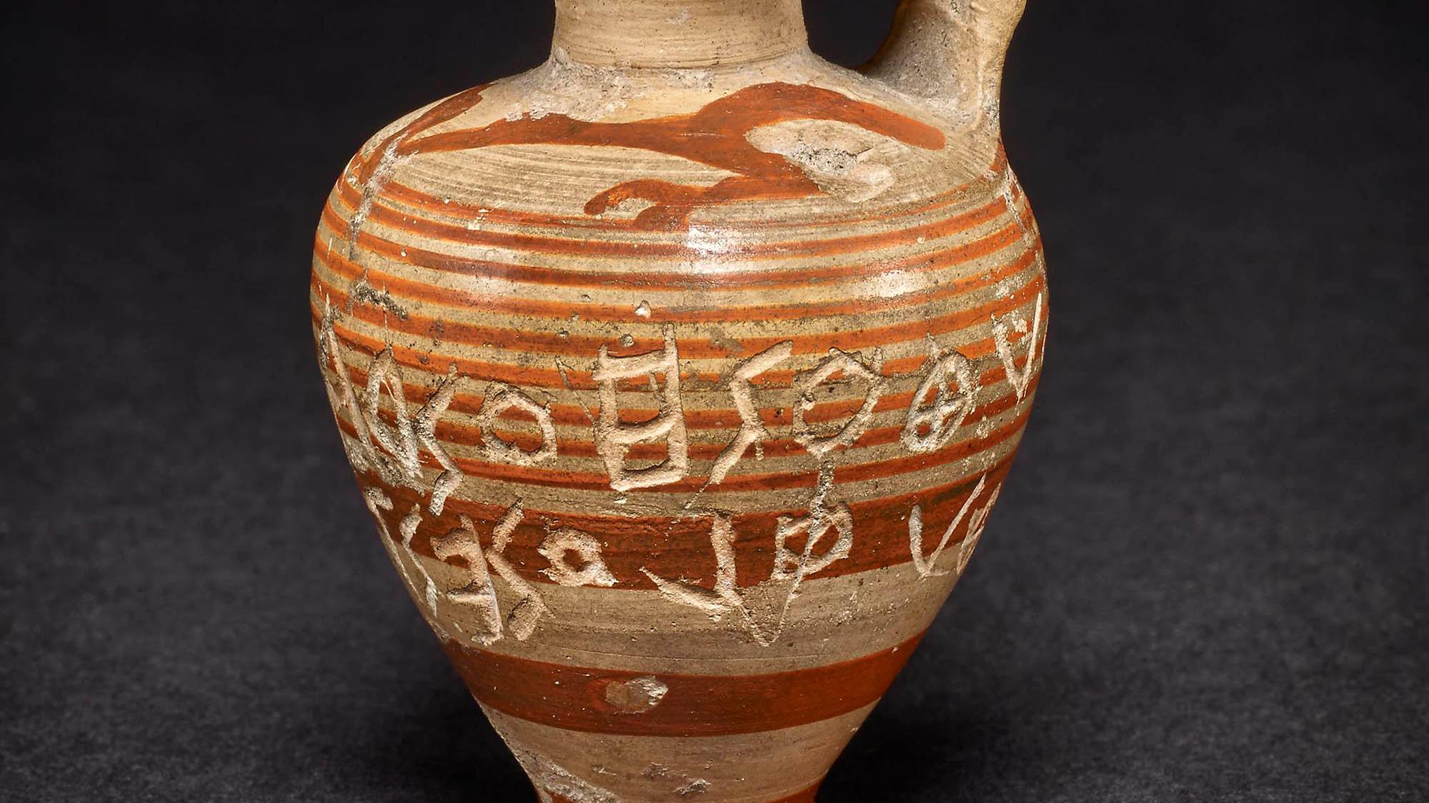 An ancient Greek clay jug with red stripes and inscriptions against a dark background.