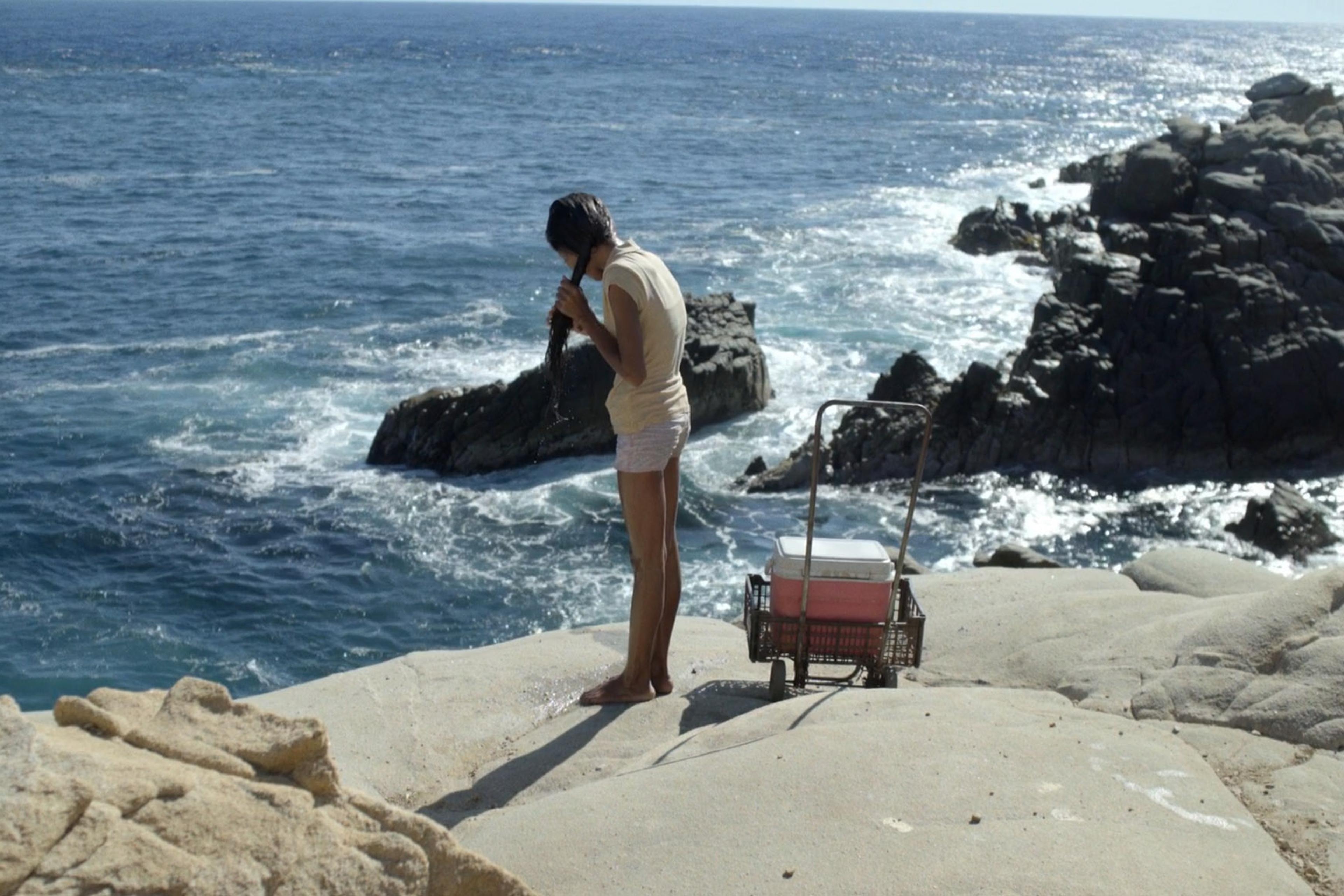 Girl standing on rocky coastline with a cooler in a cart, looking out at the ocean waves.