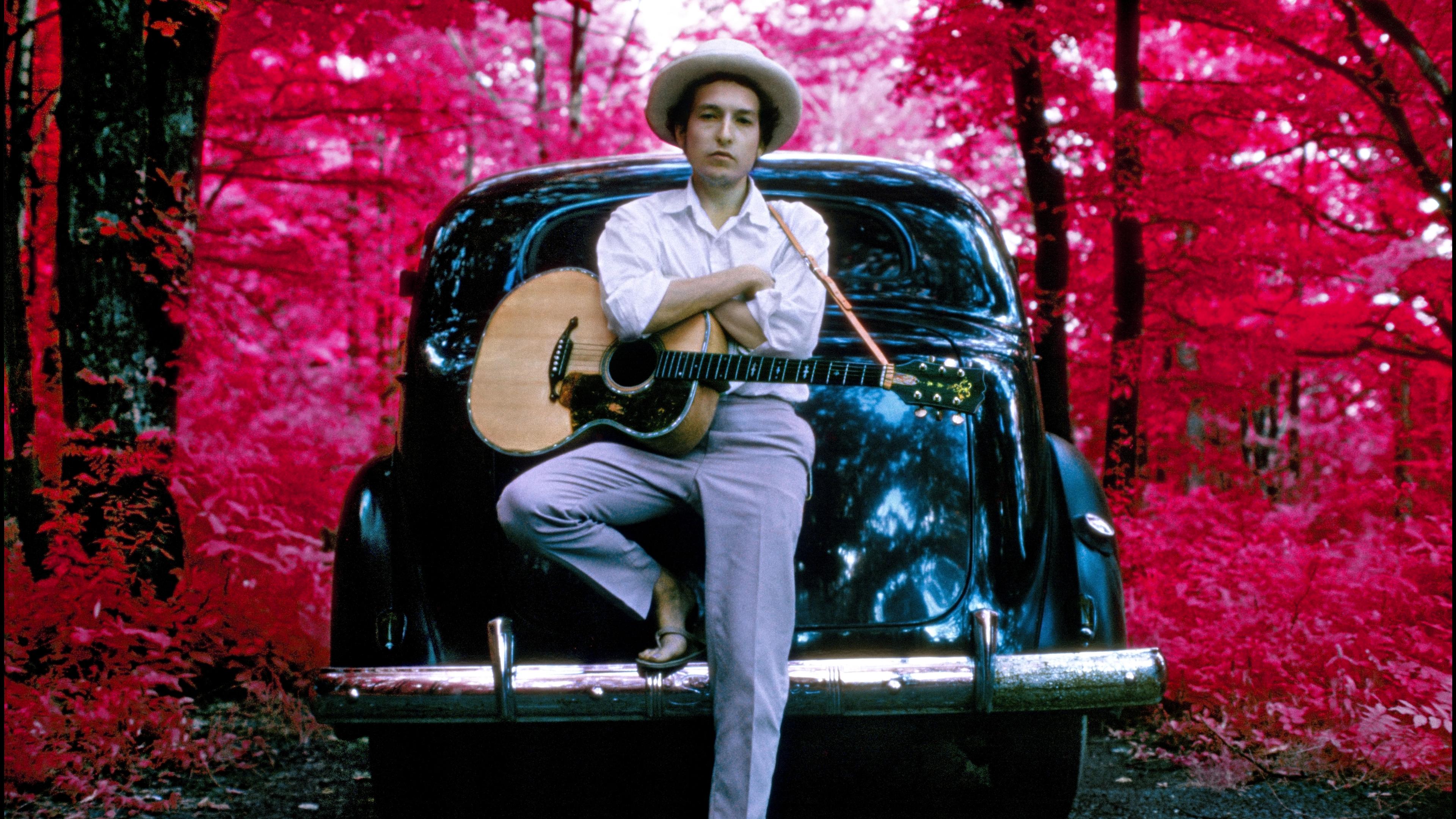 Photo of a person with a guitar leaning on a car in a forest with bright red foliage wearing a white shirt and hat.