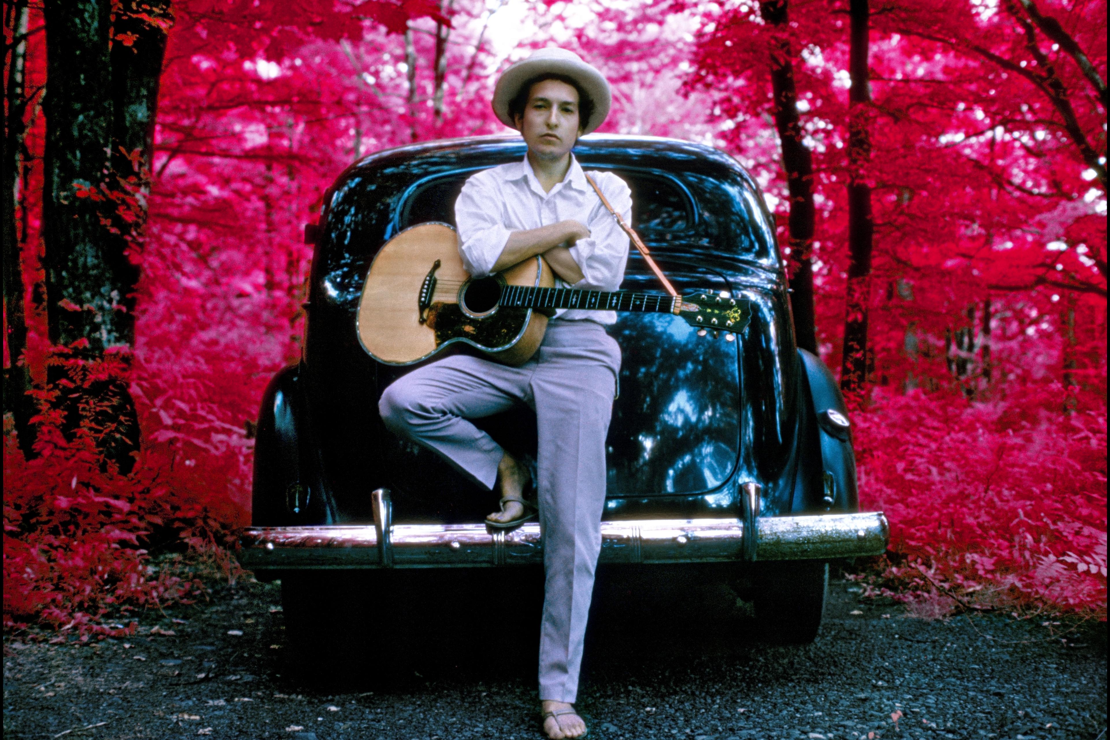 Photo of a person with a guitar leaning on a car in a forest with bright red foliage wearing a white shirt and hat.