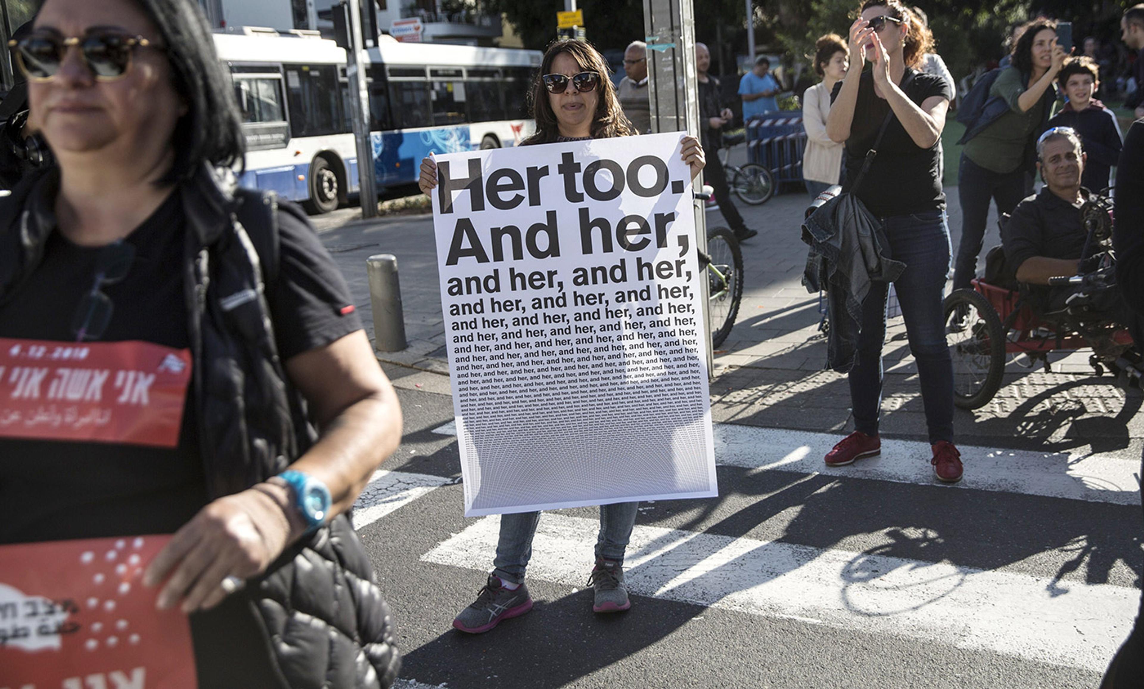 A woman holding a sign that reads “Her too. And her, and her, and her,” surrounded by people, some applauding.