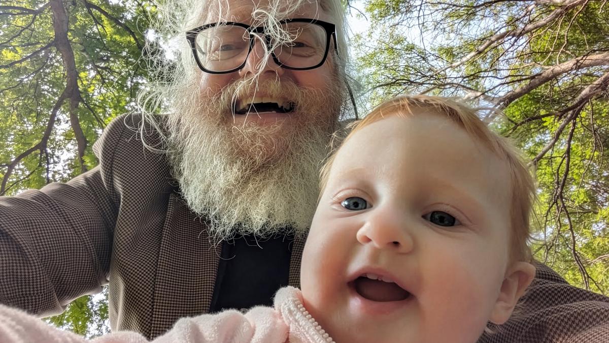 An older man with a beard and glasses and a baby smiling outdoors with trees in the background.