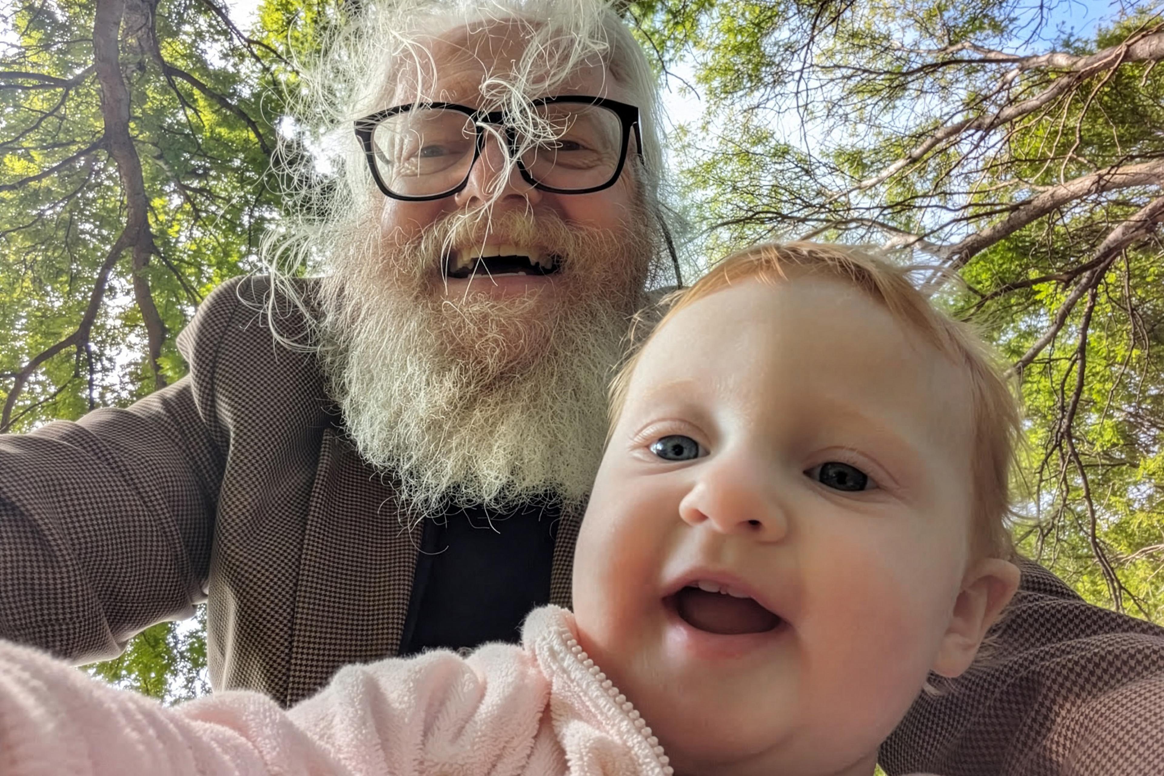 An older man with a beard and glasses and a baby smiling outdoors with trees in the background.